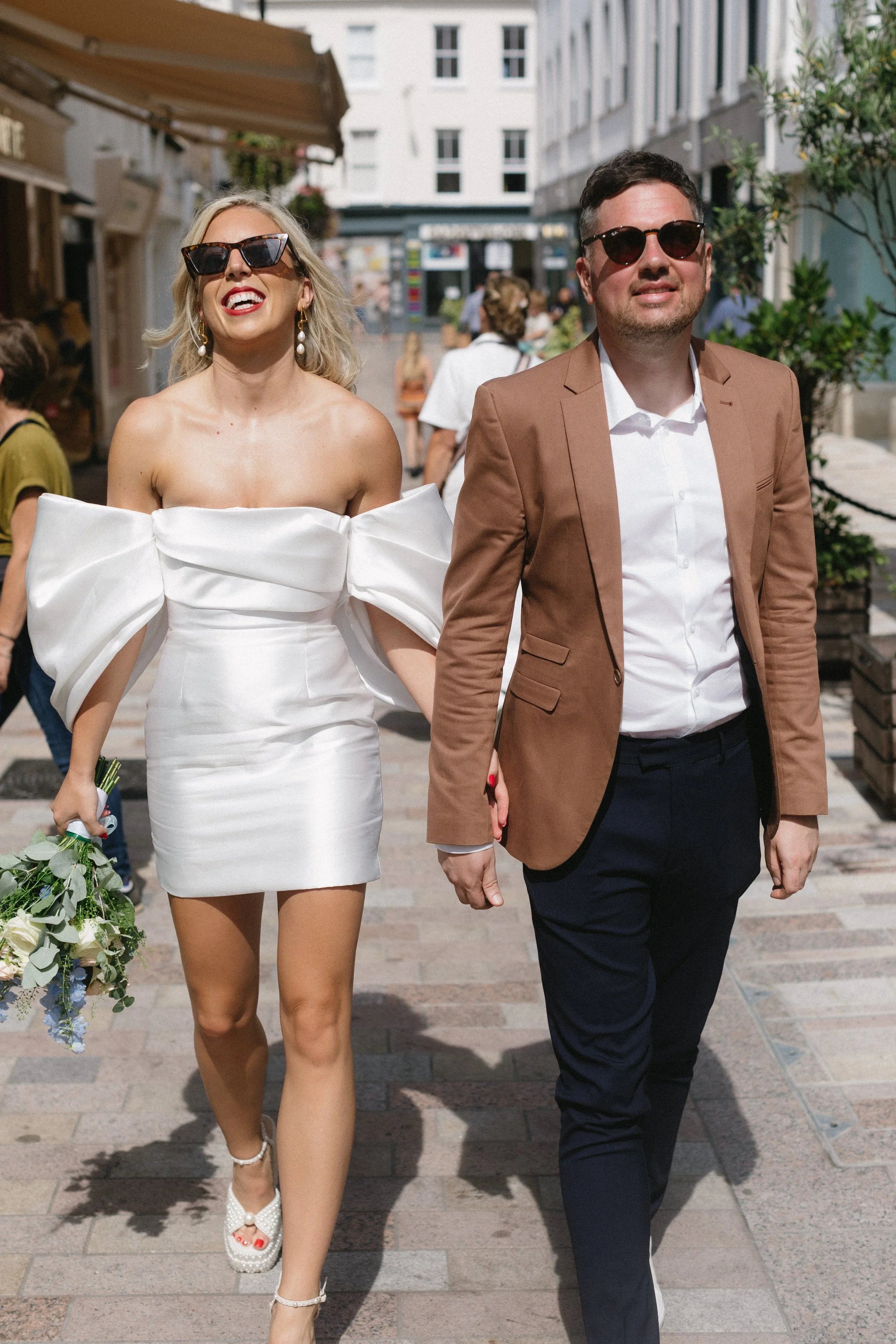 A couple walking hand in hand outdoors in a city street during daytime. The woman is in a white off-shoulder dress with large bows on the sleeves, white heels, and large sunglasses, holding a bouquet of flowers. The man is in a brown blazer, white sh