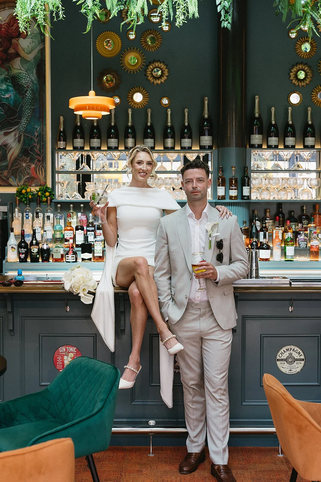 A woman in a white dress and heels sitting on a bar counter with one leg crossed and holding a drink, standing next to a man in a light beige suit with sunglasses hanging from his shirt collar, both at a bar with bottles of alcohol and decorative wal