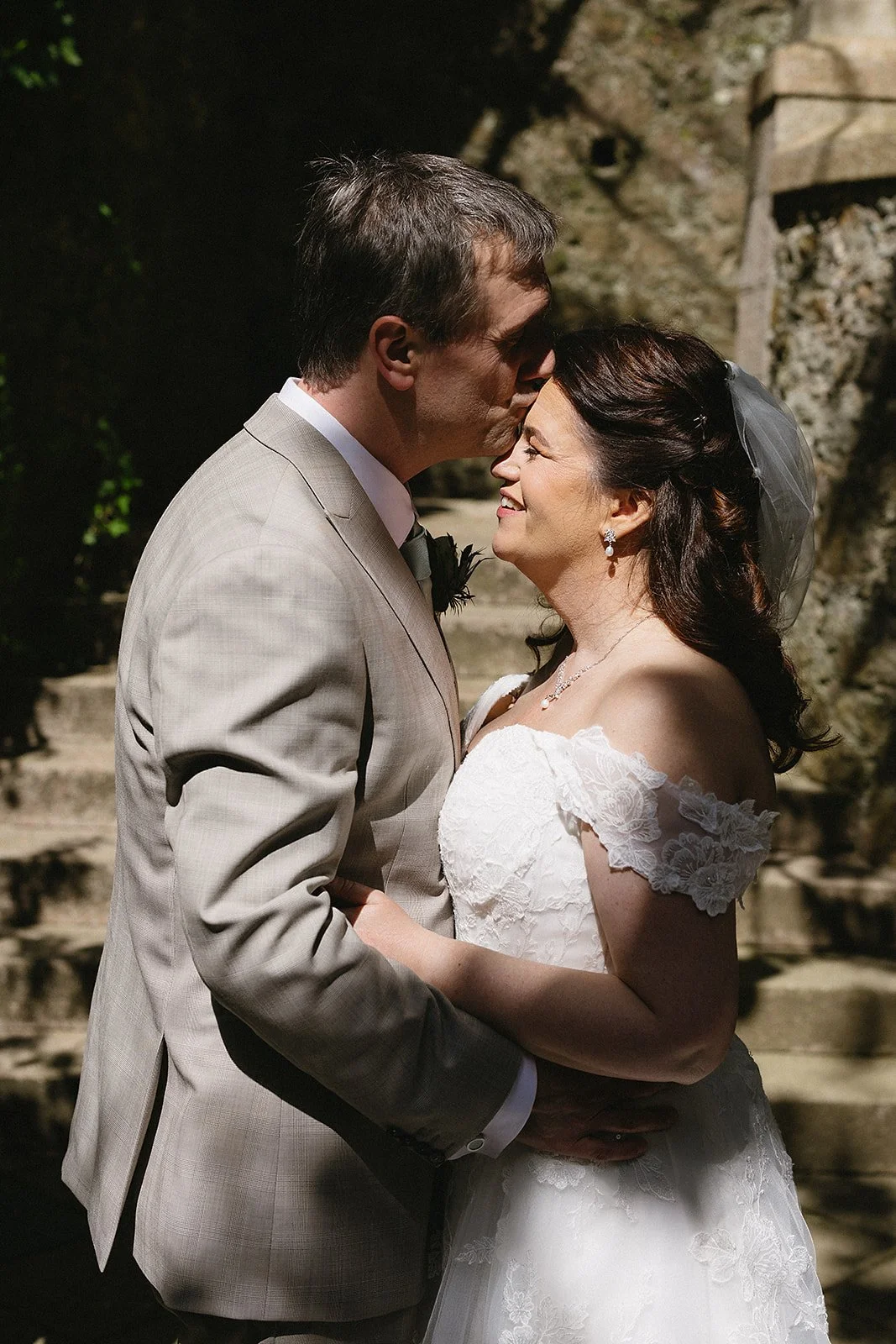 A bride and groom sharing a kiss on their wedding day, outdoors in sunlight, with stone steps in the background.