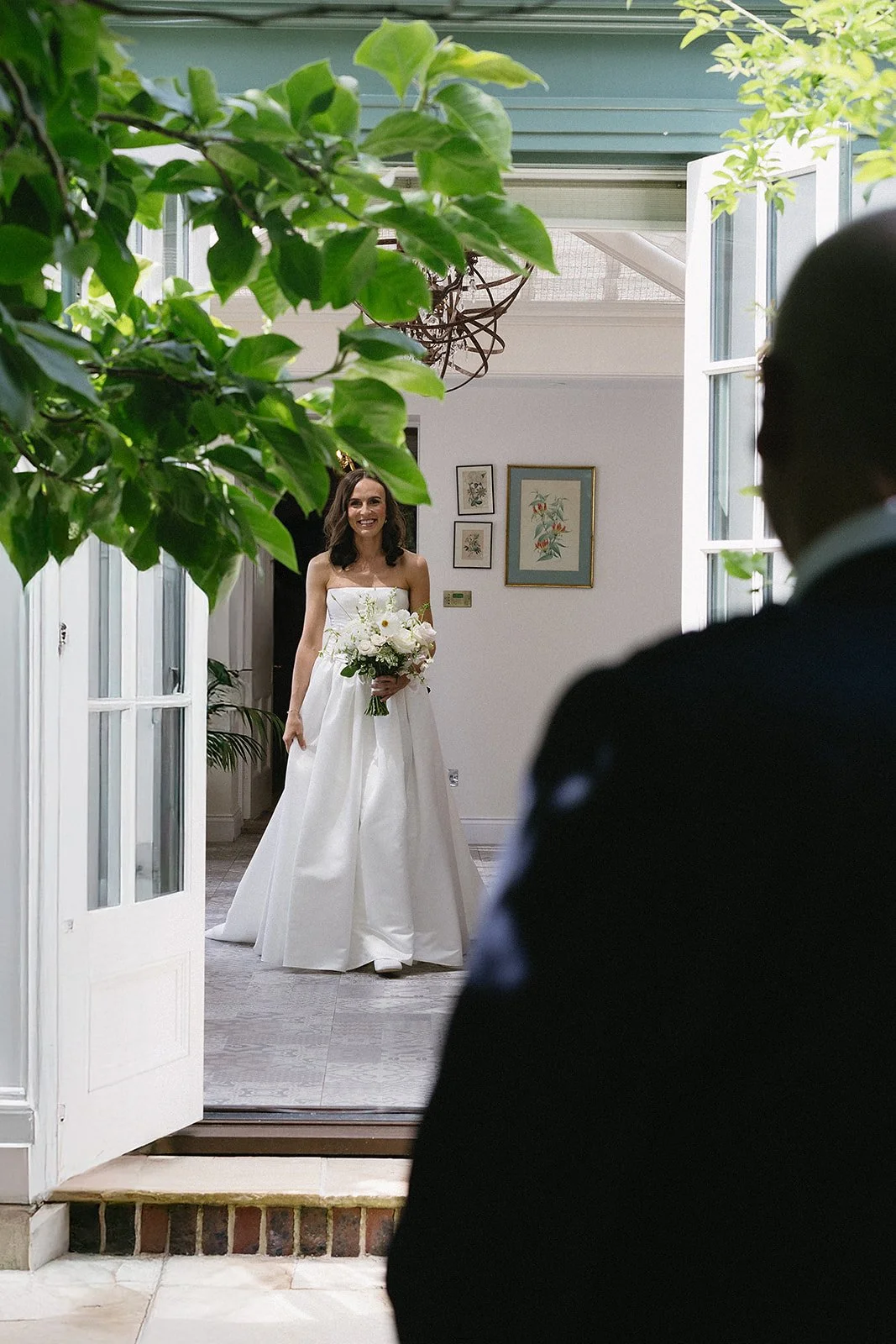 A woman in a wedding dress holding a bouquet of white flowers, smiling, standing inside a house, framed wall art, open door, and a man in a suit in the foreground.