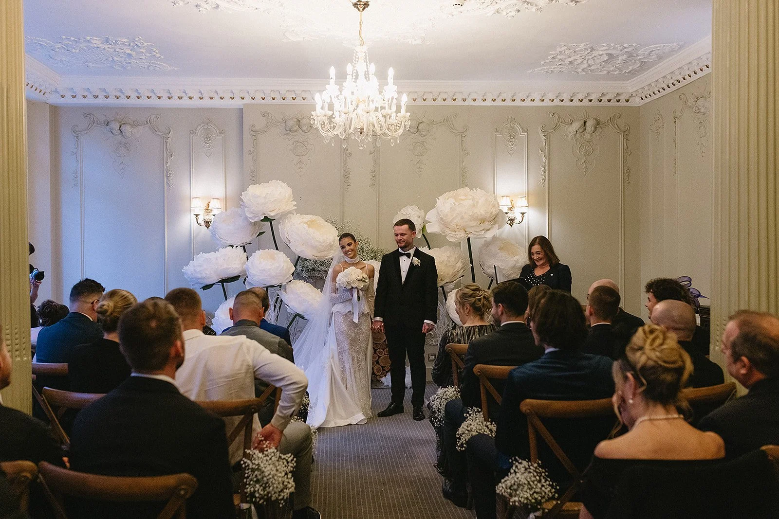 Wedding ceremony with bride and groom standing in front of guests inside a decorated room with large white flower backdrop.