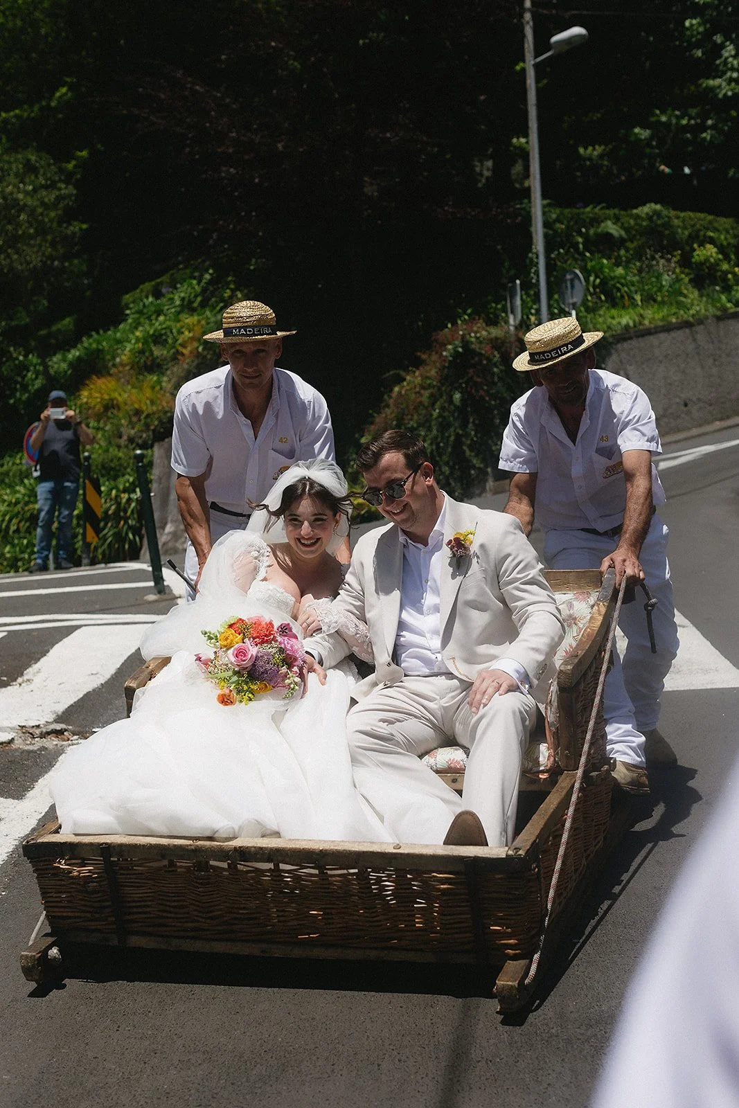 A bride and groom ride in a wooden carriage through a street, accompanied by two men in white shirts and straw hats. The bride is holding a colorful bouquet and smiling, while the groom is dressed in a light-colored suit and sunglasses, also smiling. The background shows greenery and a person taking a photo.