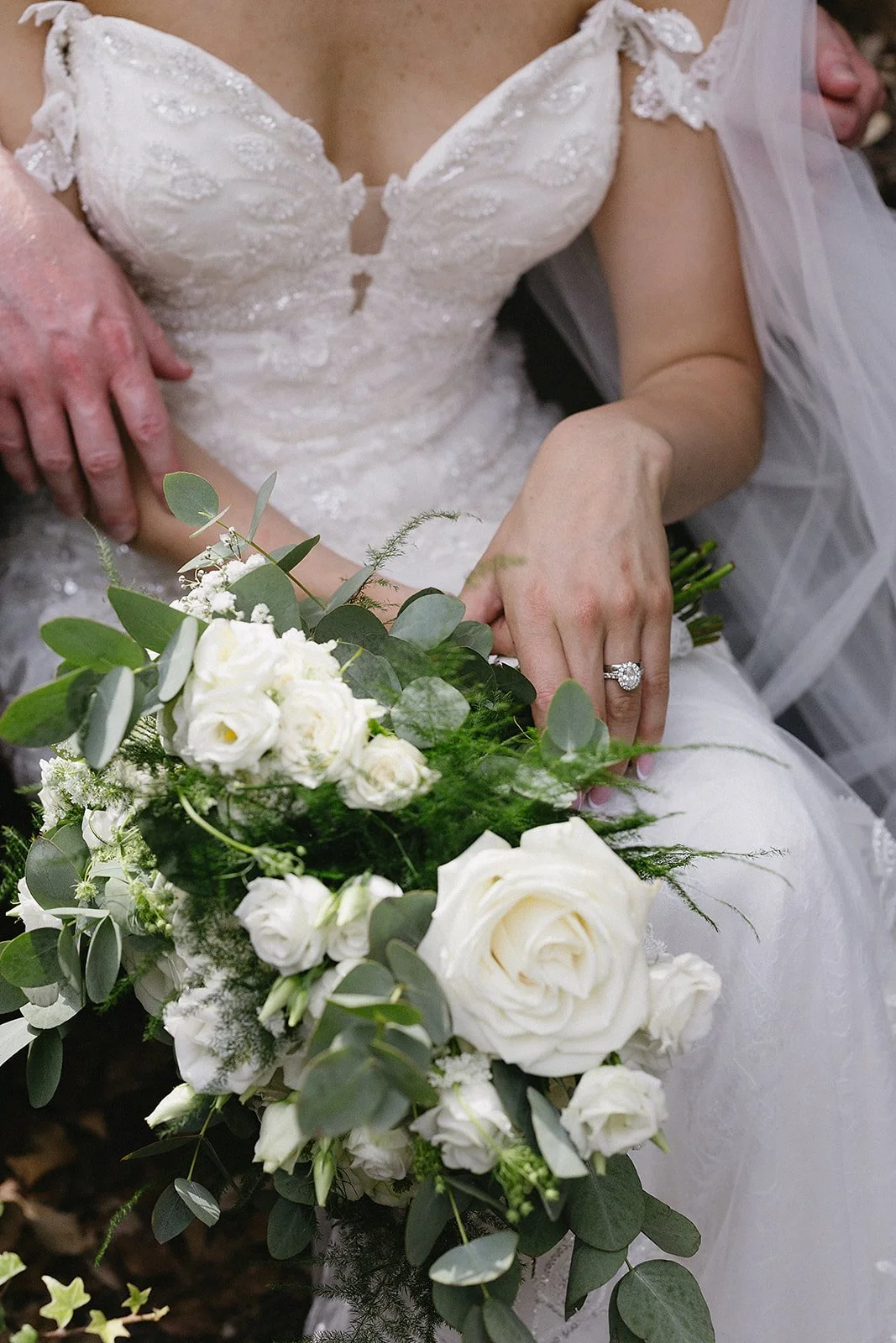 Close-up of a bride in a white lace wedding dress holding a bouquet of white roses and greenery, showing her engagement ring.