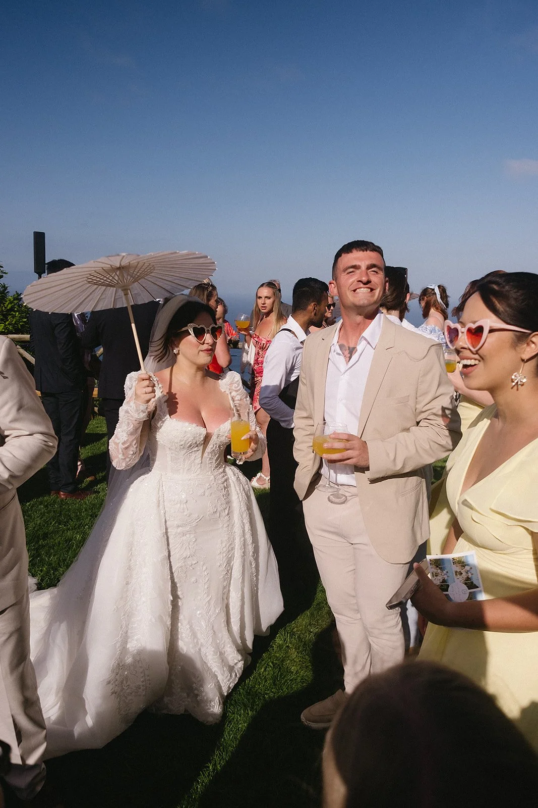 A wedding celebration outdoors with guests enjoying drinks on a sunny day. The bride in a white gown and sunglasses holds a parasol and a drink, while the groom in a beige suit and sunglasses also holds a drink. Many guests are in the background, some with drinks and summer attire.
