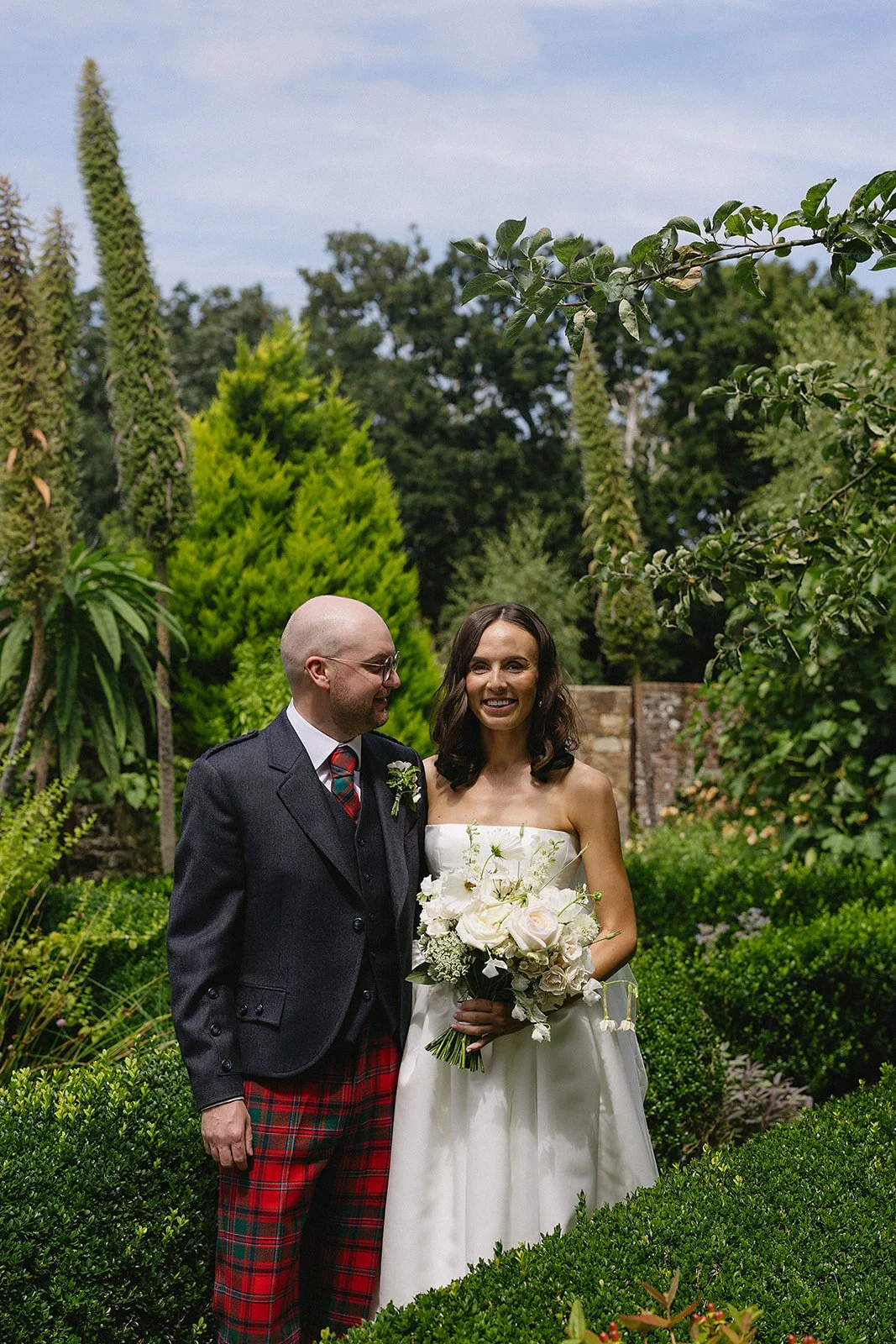 A newly married couple standing in a lush garden, the bride holding a bouquet of white flowers and wearing a strapless white wedding dress, while the groom is dressed in a dark jacket, tartan trousers, and a red tartan tie, both smiling.