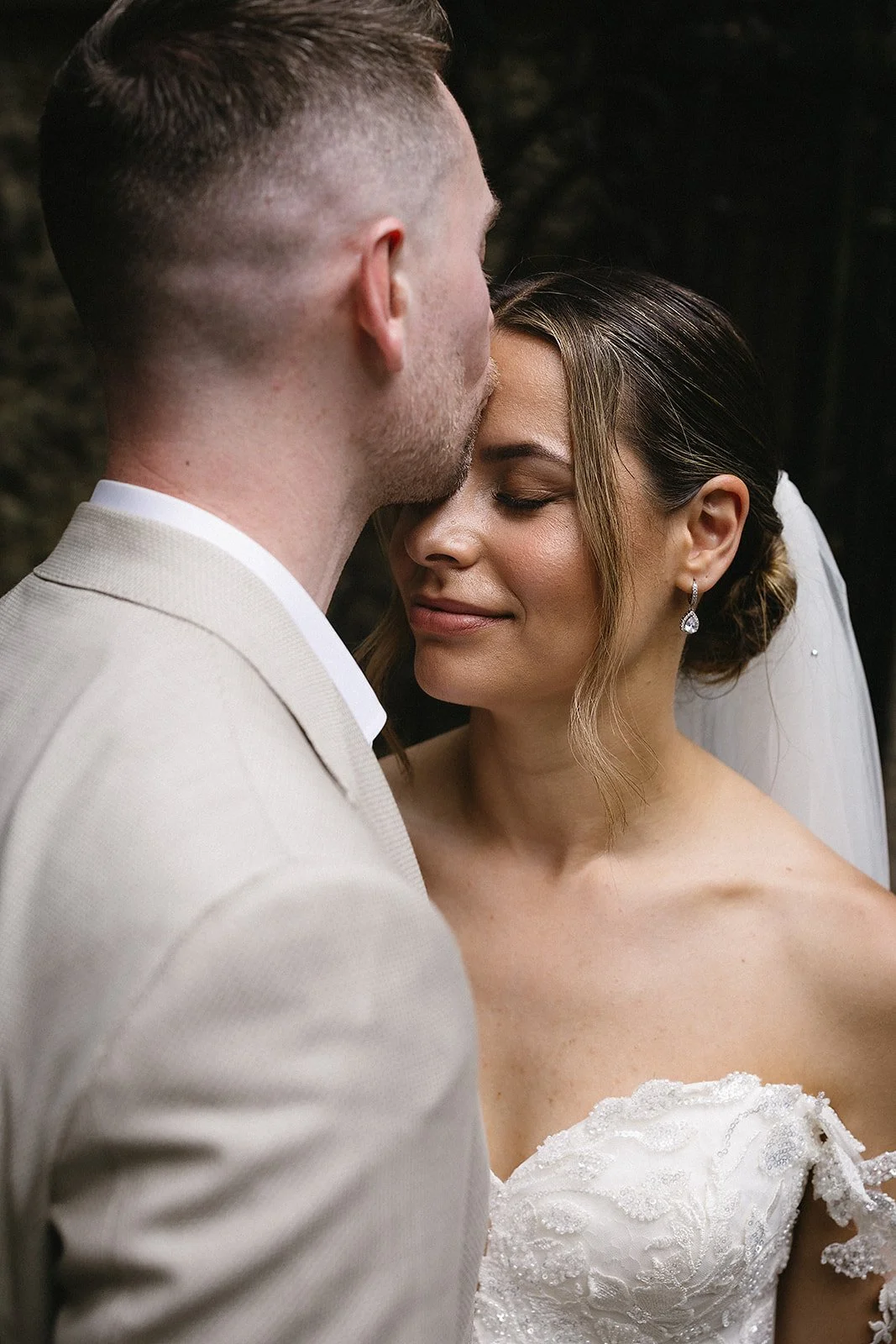 A bride and groom sharing an intimate moment, with the groom kissing the bride's forehead as she smiles softly with her eyes closed.