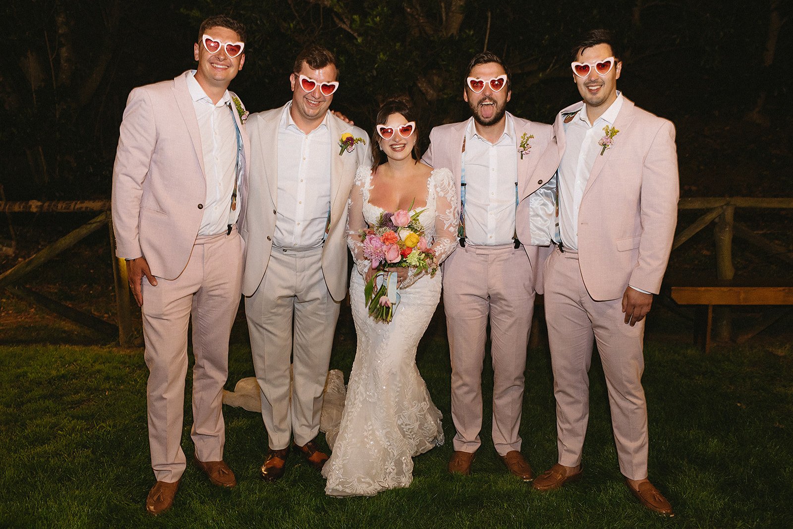 A bride in a white lace wedding dress holding a colorful bouquet, surrounded by five men wearing matching pink suits, all wearing heart-shaped sunglasses, standing outdoors in front of a wooden fence at night.