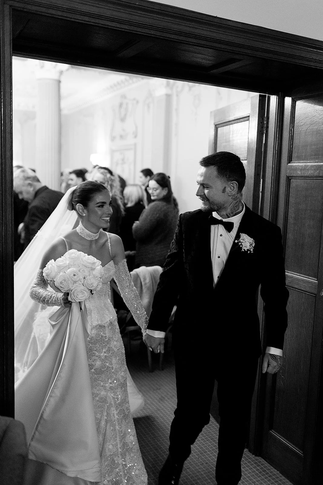 A black and white photo of a bride and groom holding hands, smiling, as they walk through a doorway inside a venue, with guests in the background.
