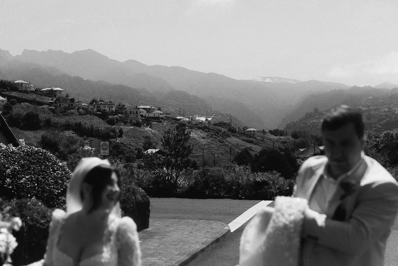A black-and-white photo of a man and woman in foreground, with a scenic hilly landscape and houses in the background.