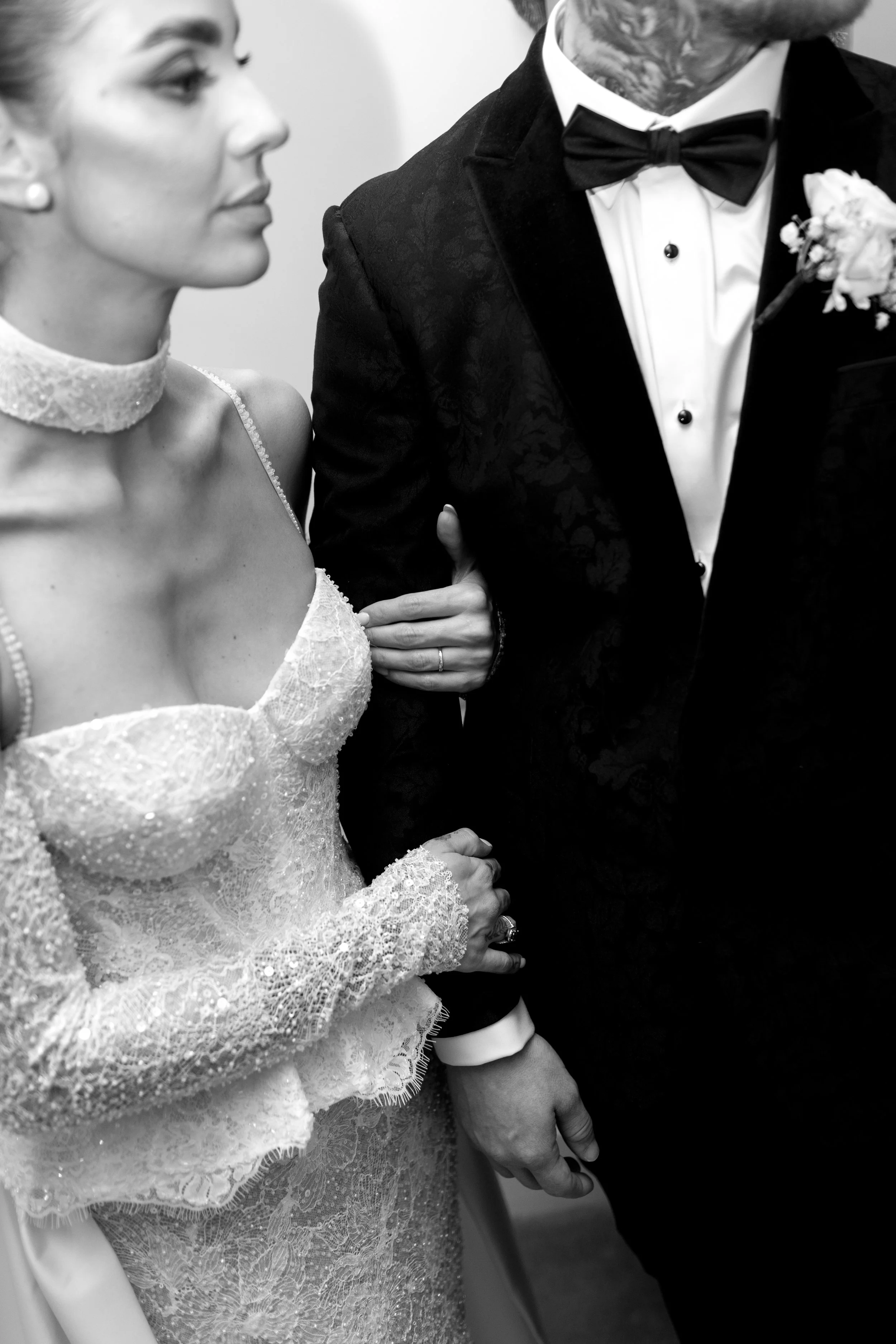 Black and white photo of a woman in a lace wedding dress and a man in a tuxedo with a bowtie, holding hands during a wedding ceremony.