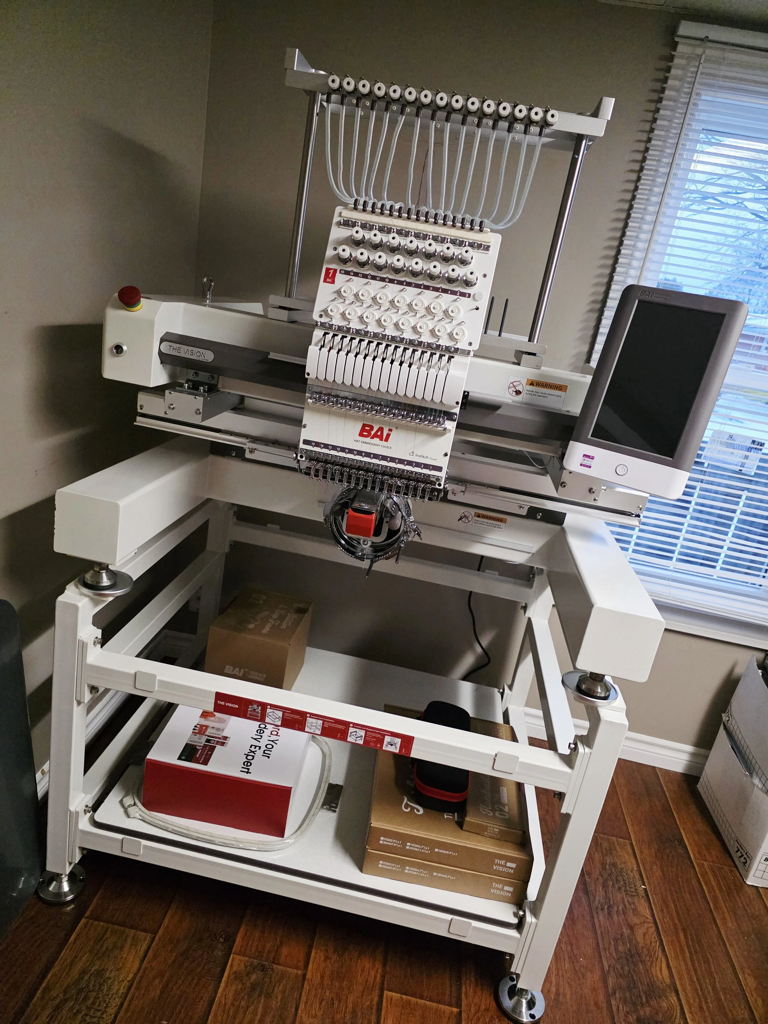 A computerized embroidery sewing machine with multiple thread spools, a monitor screen, and a small worktable with boxes and equipment underneath.
