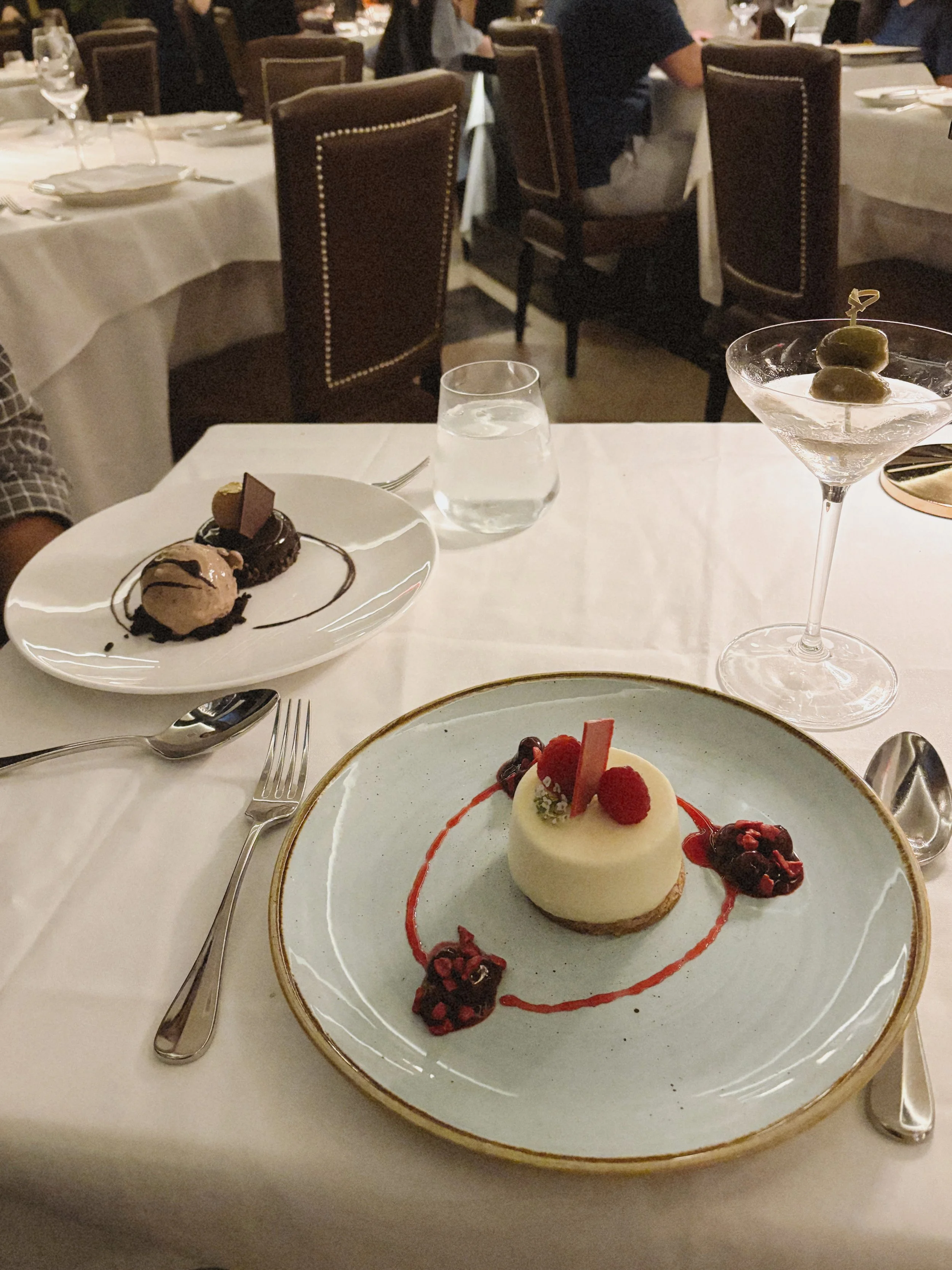 A dessert plate with a small round cheesecake topped with raspberries, served with berry compote, garnished with decorative sauce, on a large white plate with a gold rim, on a white tablecloth in a restaurant setting.