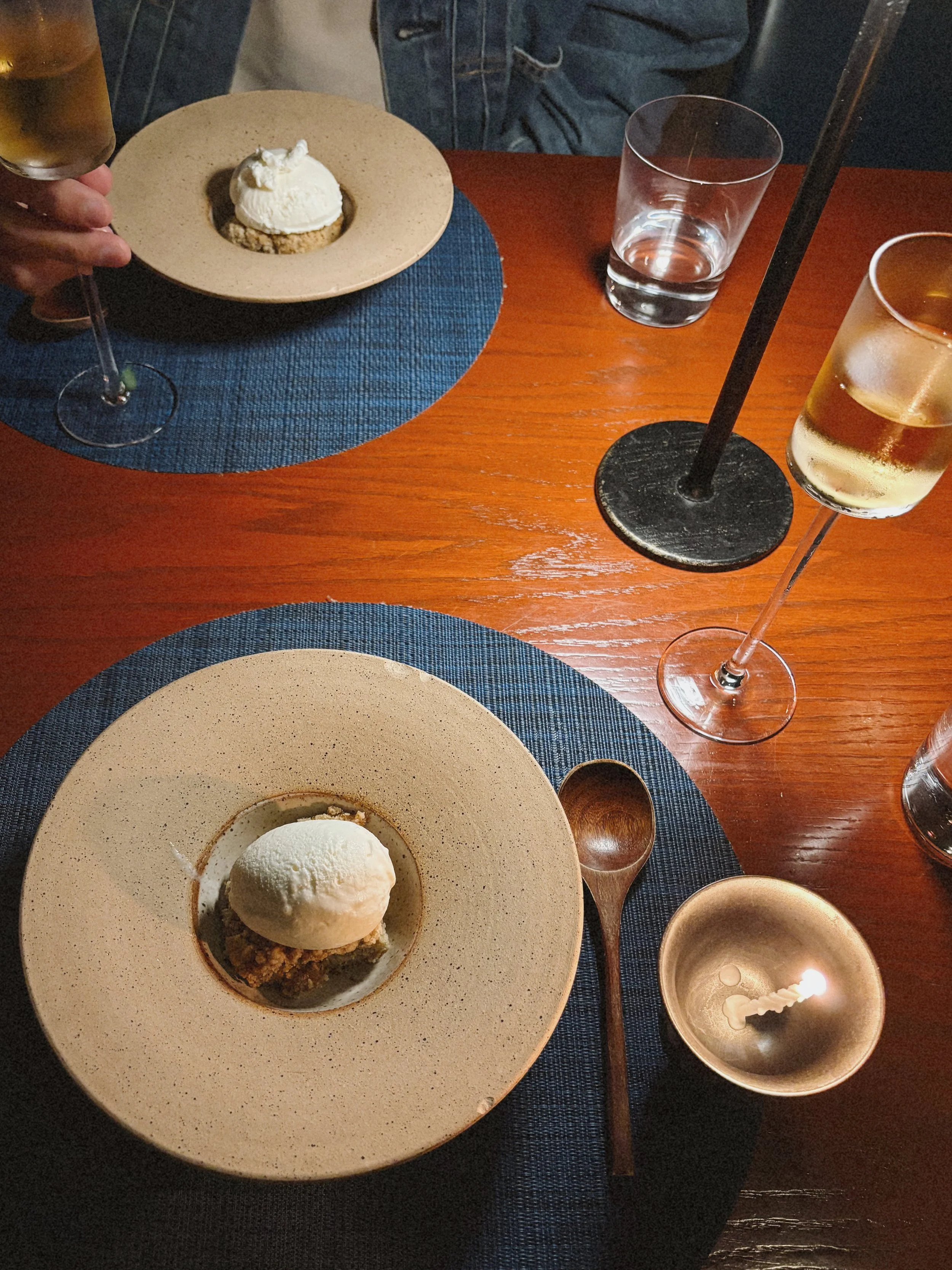 Dessert plate with a scoop of vanilla ice cream and a crumbly base, seen on a table with blue placemats, along with a glass of water, a champagne flute, and a lit candle.