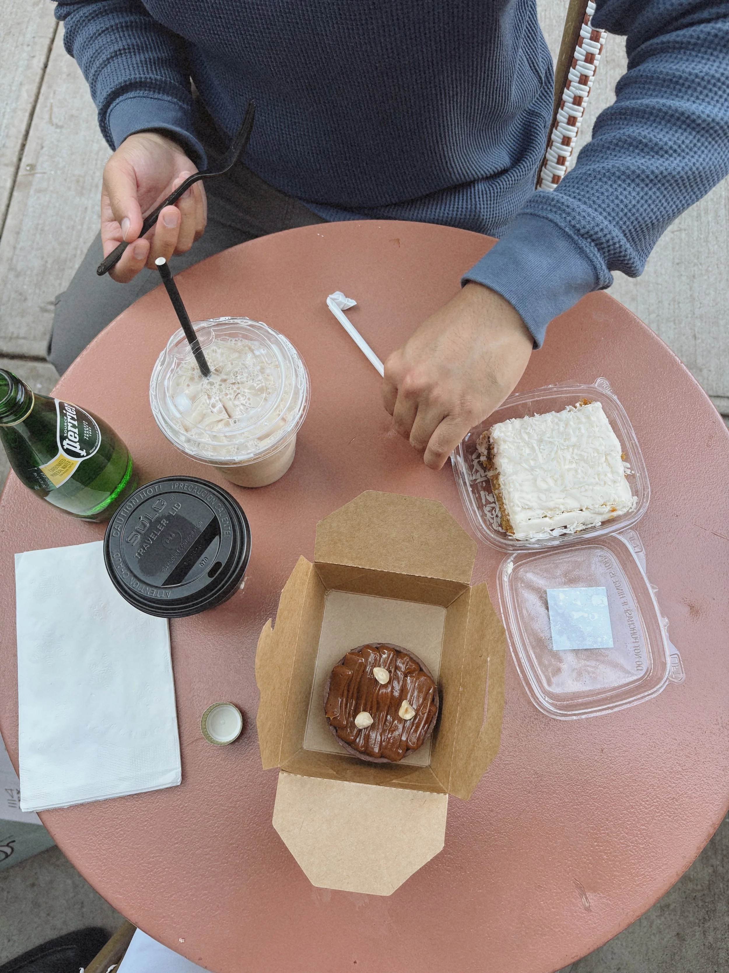 Top-down view of a person at a pink table with a bakery box containing a round chocolate cake, a plastic container with a white frosted cake, a plastic cup with iced beverage, a green beer bottle, a black takeout container, a paper napkin, and a small bottle of white sauce.