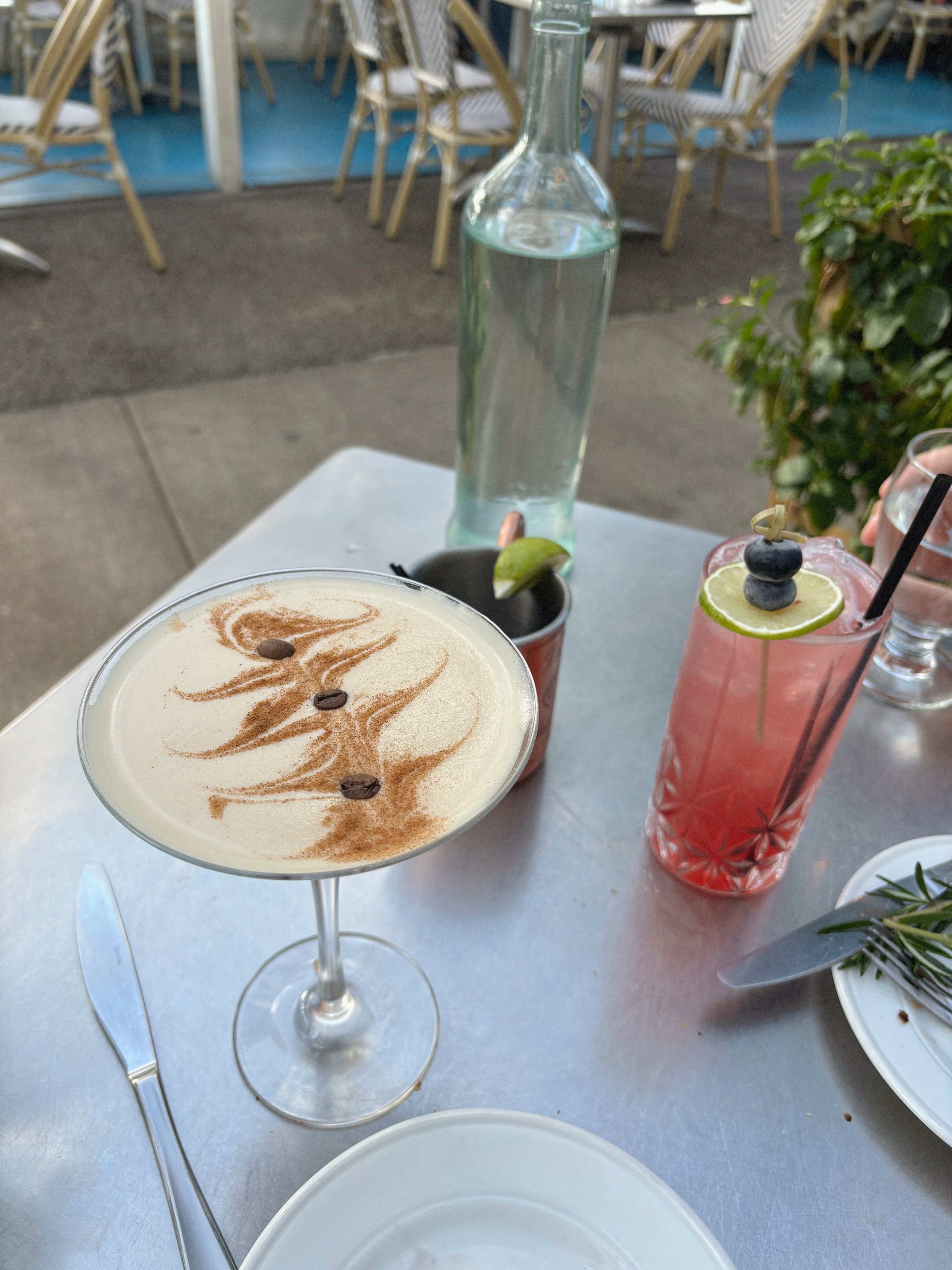 A table with a cocktail garnished with lime and blueberries, a small black cup with a lime wedge, a tall clear water bottle, and a pink drink with a lime slice and blueberries. In the background are chairs and tables at an outdoor cafe.