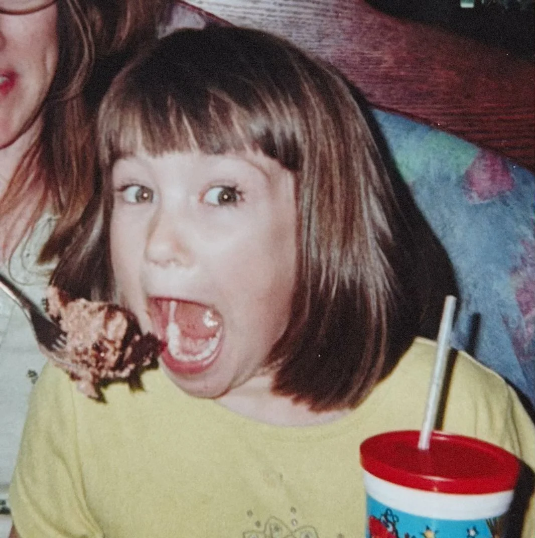A young girl with brown hair and bangs is holding a fork with a bite of cake near her open mouth, appearing excited. There is a red and white cup with a straw in front of her, and part of another person's face is visible on the left. The background shows a colorful fabric and a wooden surface.