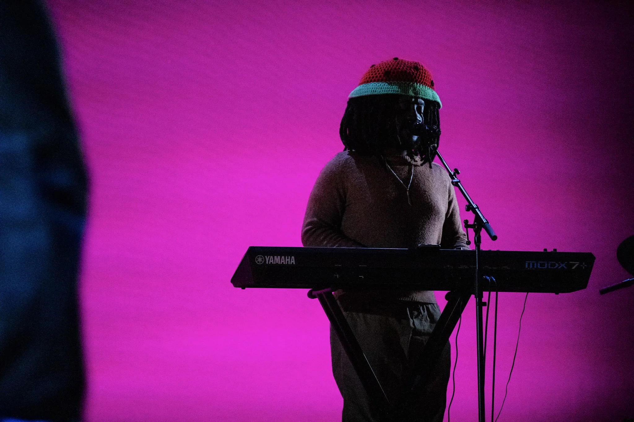 Person playing Yamaha keyboard on stage with vibrant pink background, wearing a colorful knit hat and casual clothing.