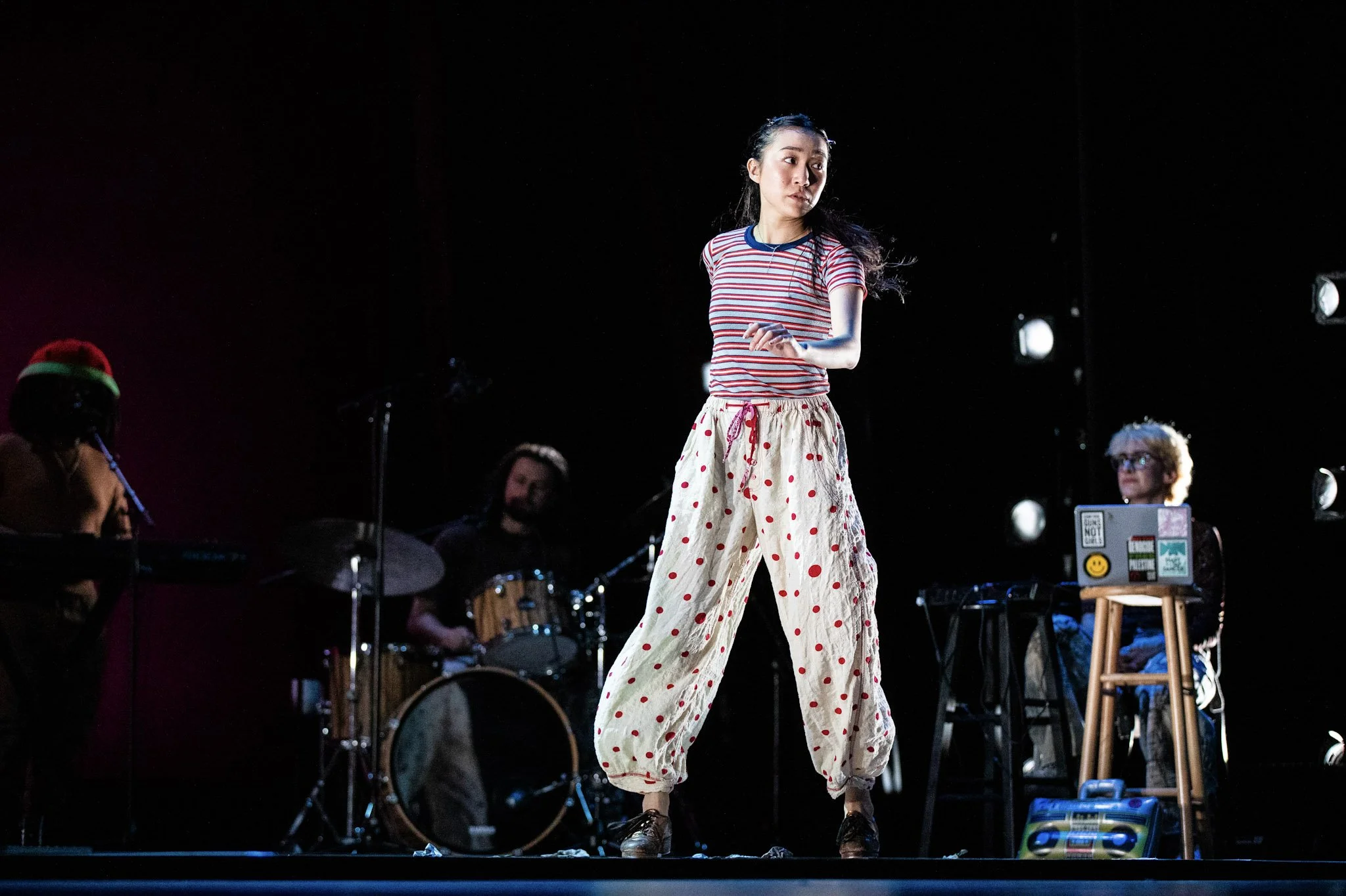 A young woman standing on stage during a performance, wearing a striped shirt and loose, polka-dot pants, with a band playing instruments behind her.
