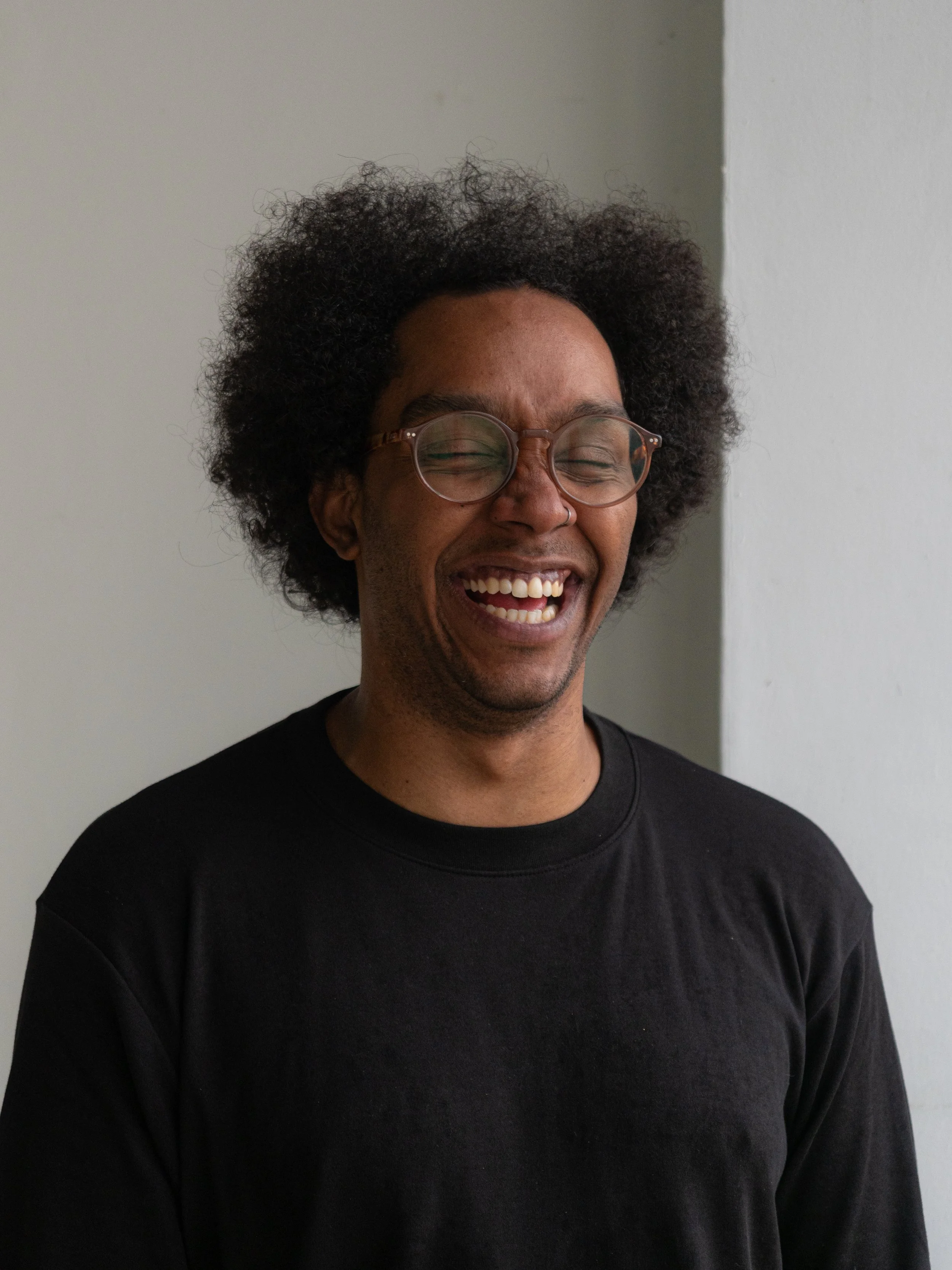 A man with curly hair and glasses laughing with eyes closed, wearing a black shirt, standing against a light-colored wall.