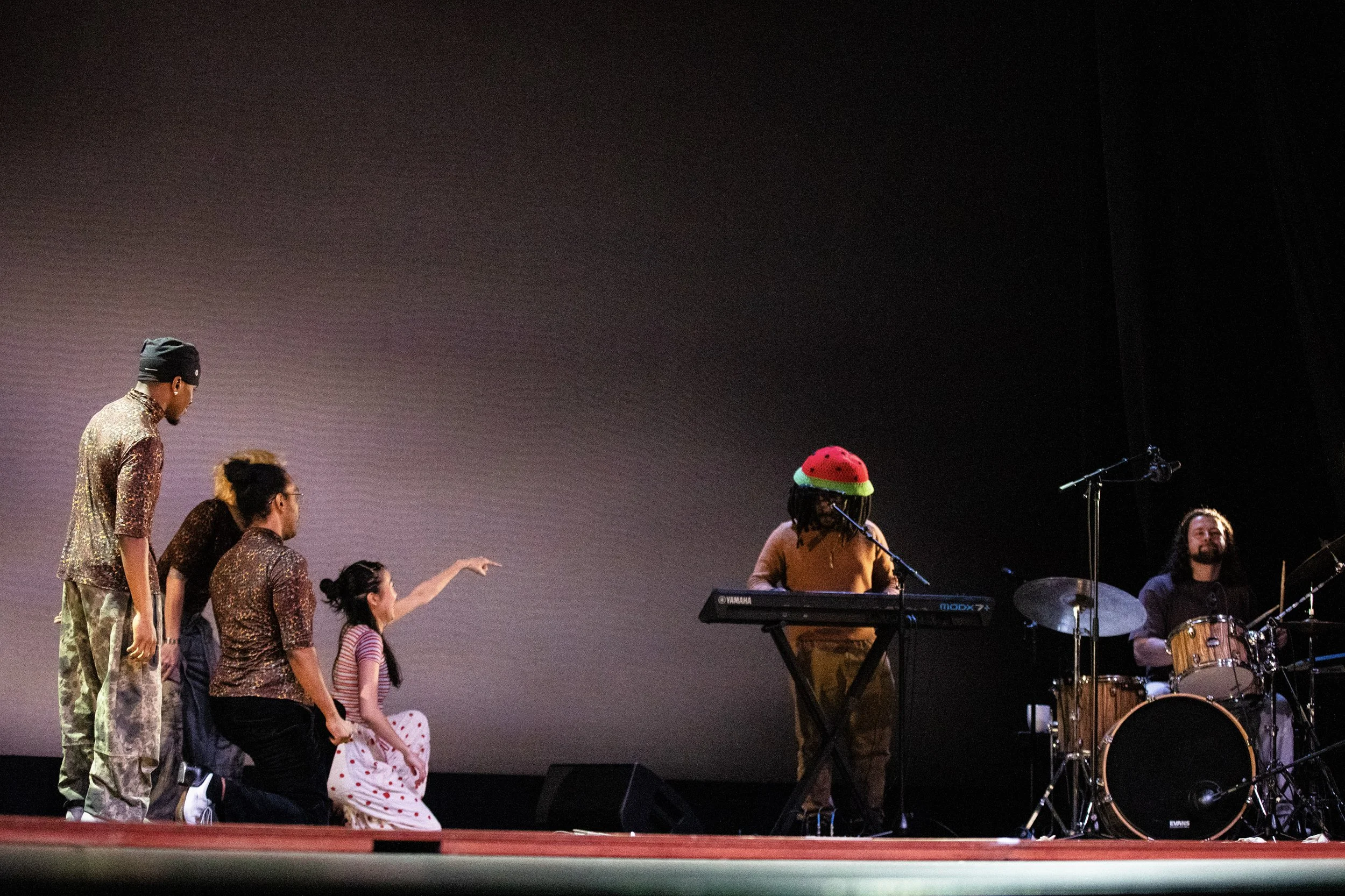 A group of five people on stage, four kneeling or standing and one sitting behind musical instruments, with two people at the keyboard and drum set on the right, a person with a red hat playing keyboard, and the others in patterned clothing, possibly rehearsing or performing.