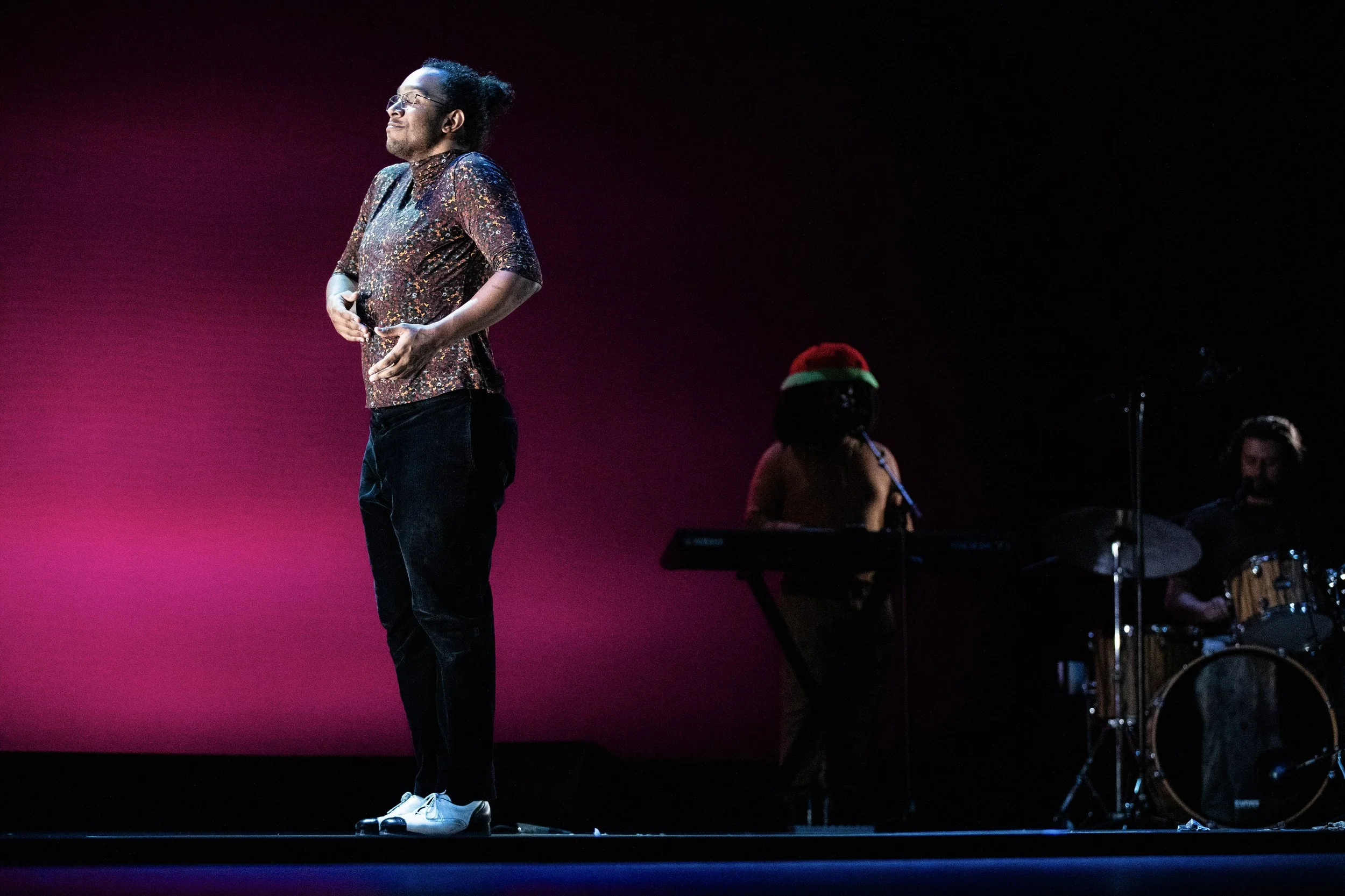 A person with glasses and curly hair stands on stage under pink and purple lighting, wearing a sequined shirt, black pants, and white shoes, with two musicians playing instruments in the background.