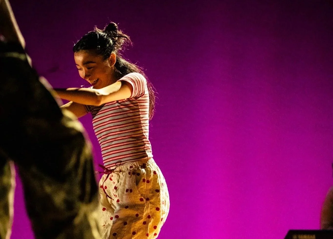 A young girl smiling and laughing, wearing a pink and white striped shirt and white pants with red polka dots, against a purple background.