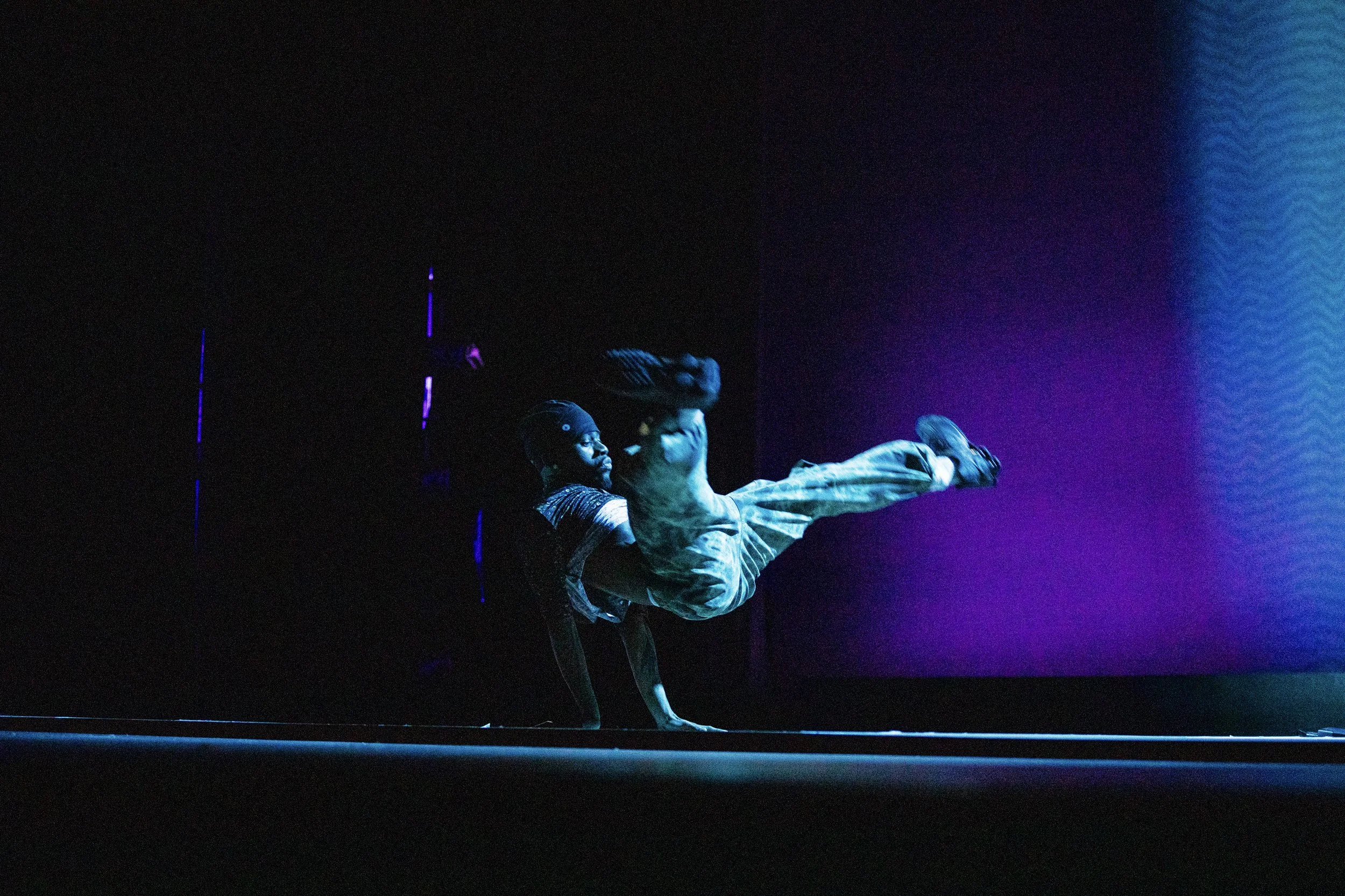 A man performing a breakdancing move on a dark stage with colorful purple and blue lighting.