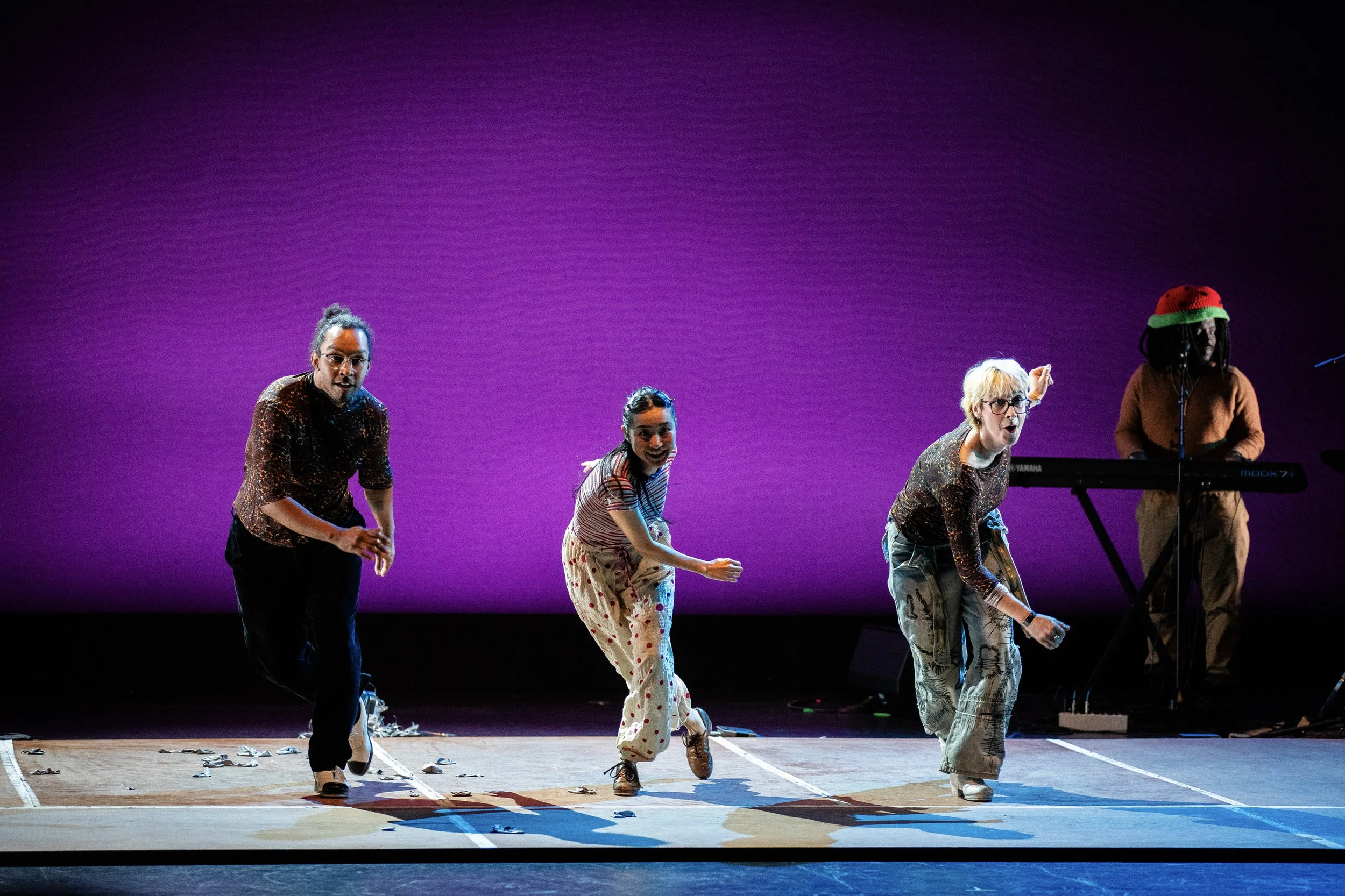 Four performers dancing and singing on stage with a purple gradient backdrop, with a keyboard player in the background wearing a red and green hat.