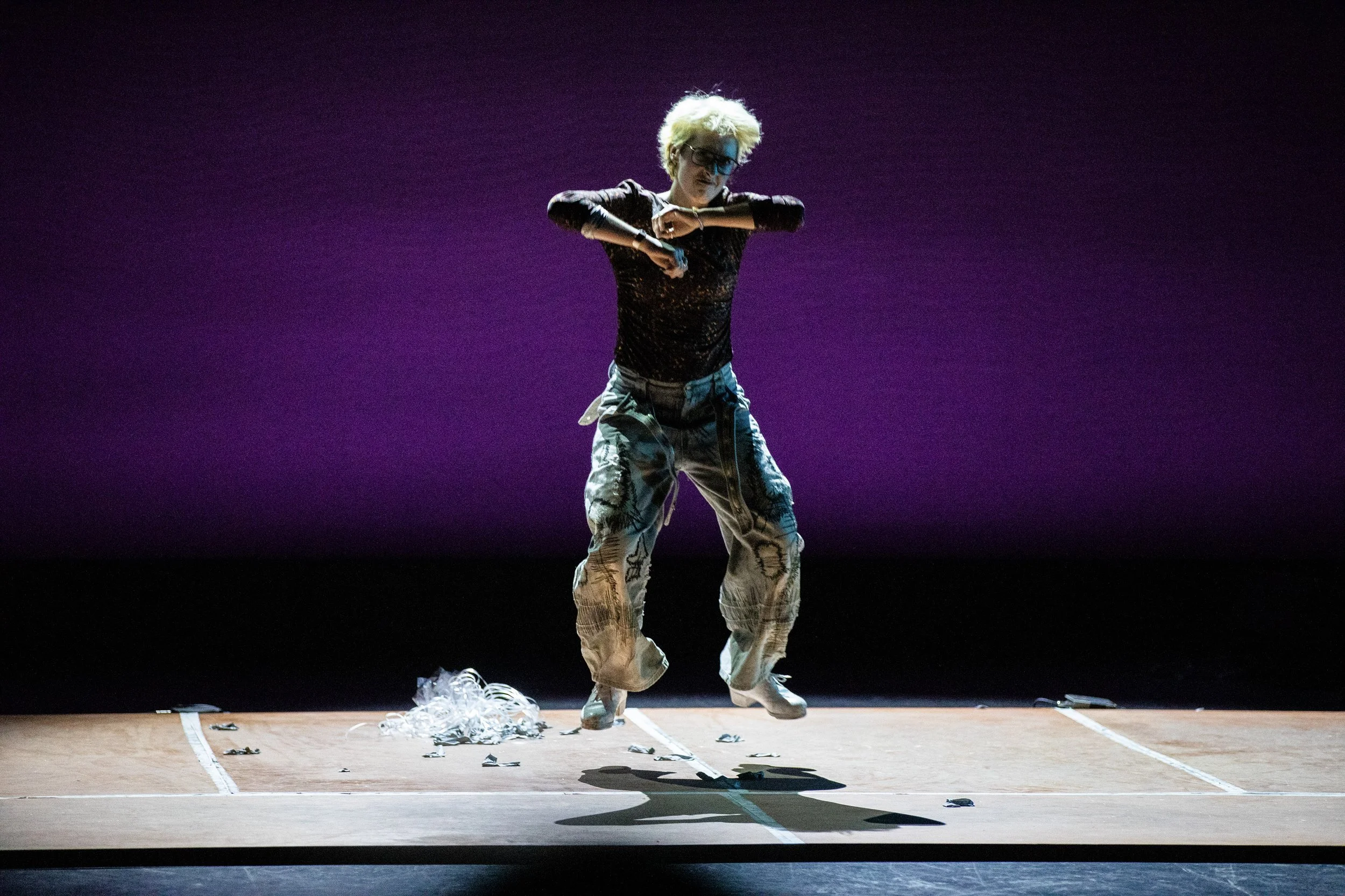 Person with blonde hair, black glasses, wearing a black sheer top and baggy cargo pants, performing on a stage with a purple background, surrounded by shredded paper on the floor.