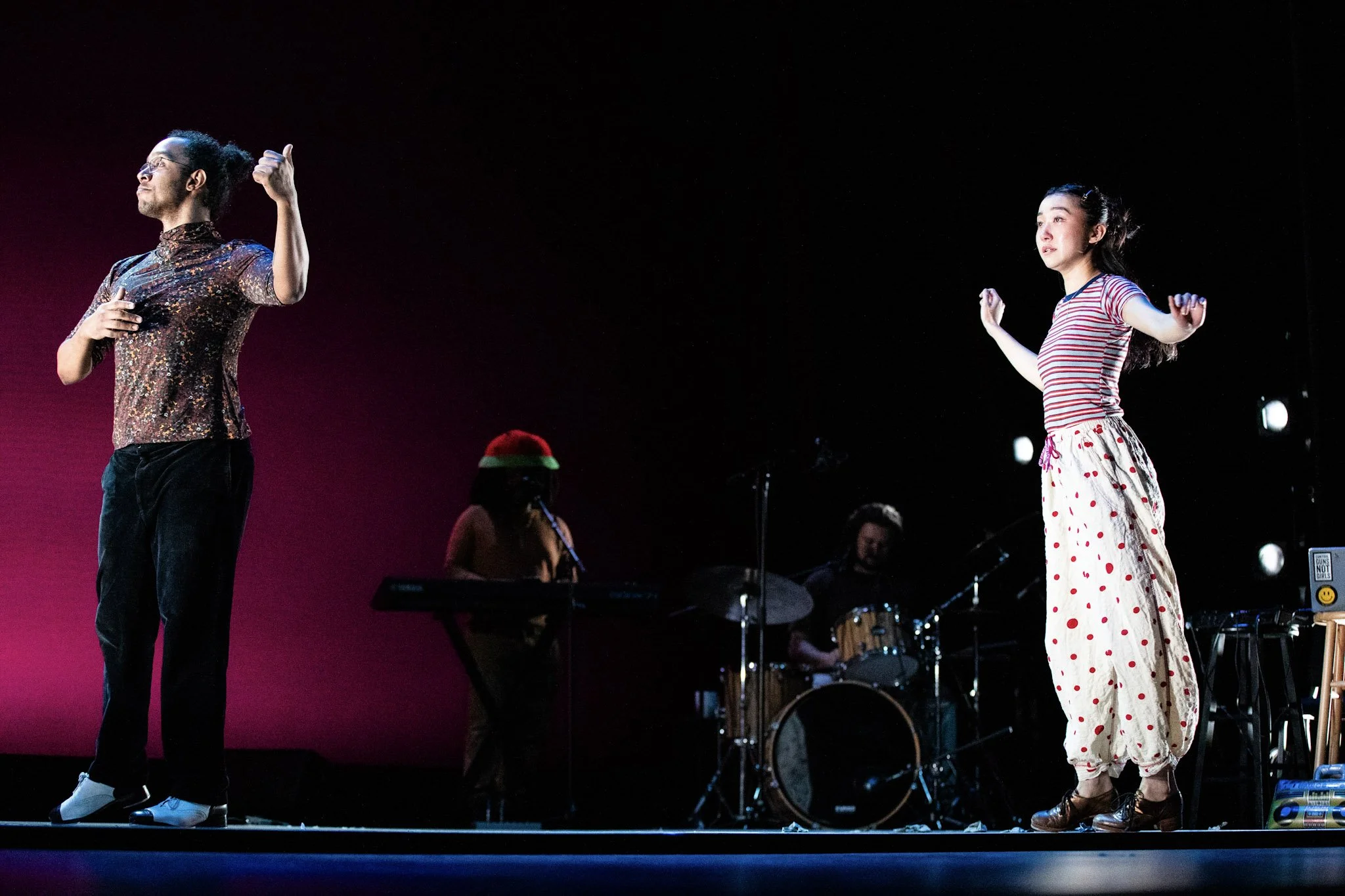 Two performers on stage, one man with glasses, curly hair, and a colorful shirt giving a thumbs up, and one woman with long dark hair, wearing striped shirt and polka dot pants, standing with arms raised. Two musicians are in the background playing instruments.