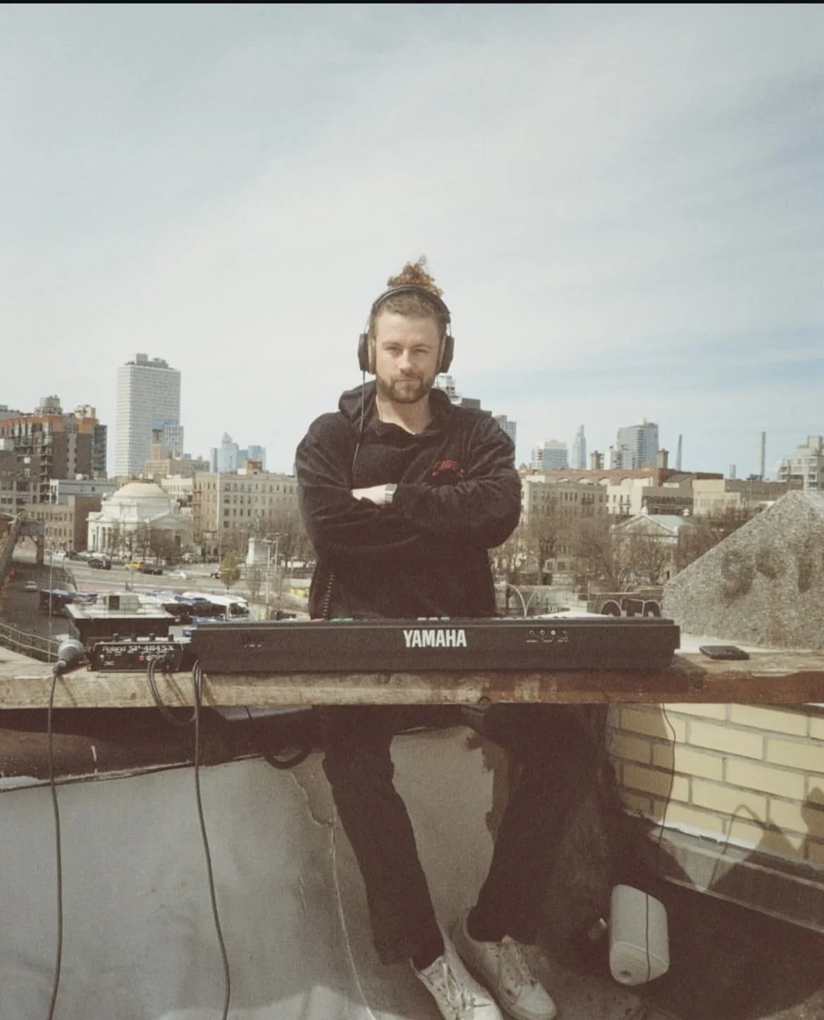 A man with a beard and long hair tied in a bun wearing headphones, standing with arms crossed behind a Yamaha keyboard on a rooftop with an urban cityscape background.