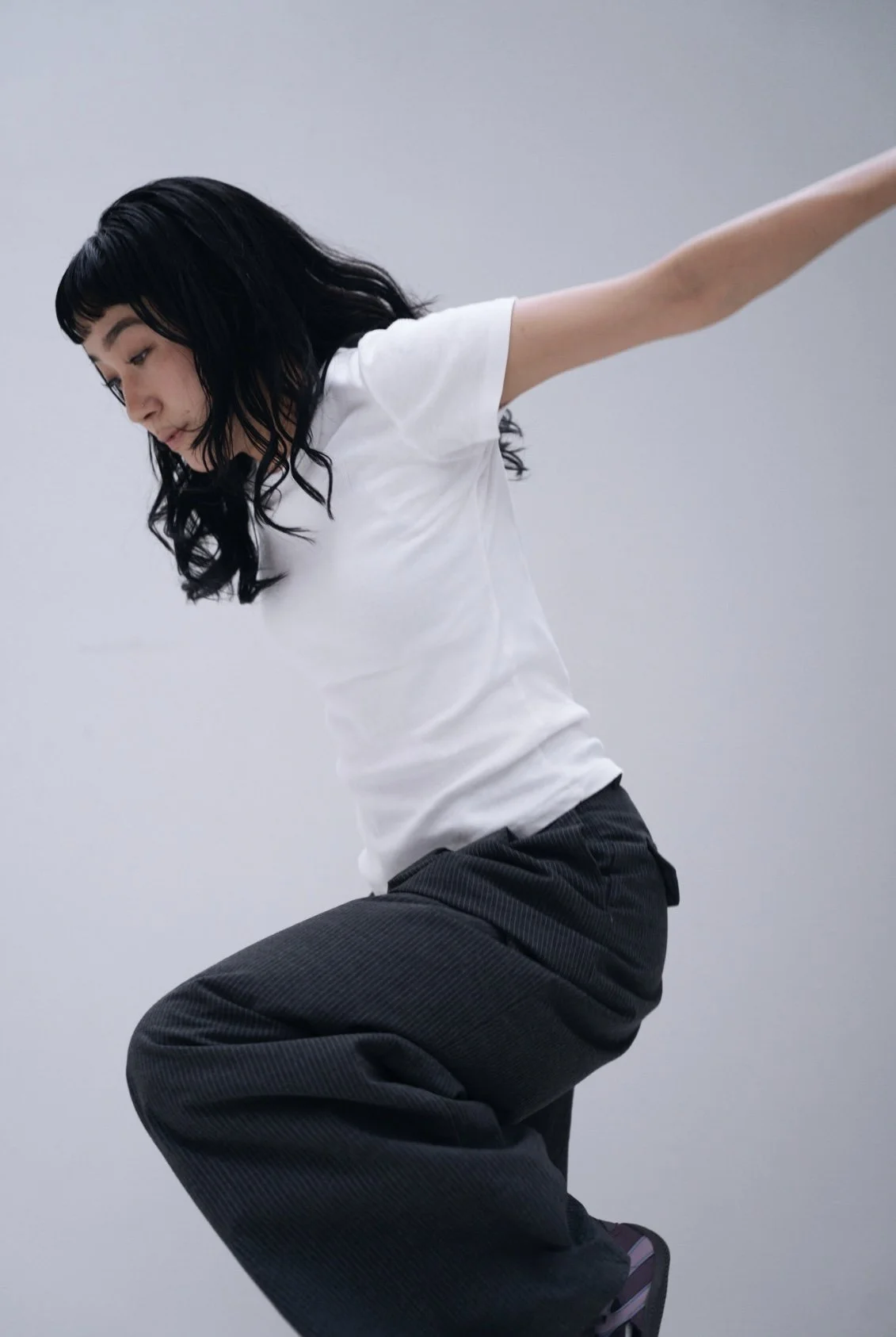 A woman with black hair wearing a white t-shirt and black pants is captured in a side view, balancing on one leg with her other leg bent and her arms extended outward, against a plain light gray background.