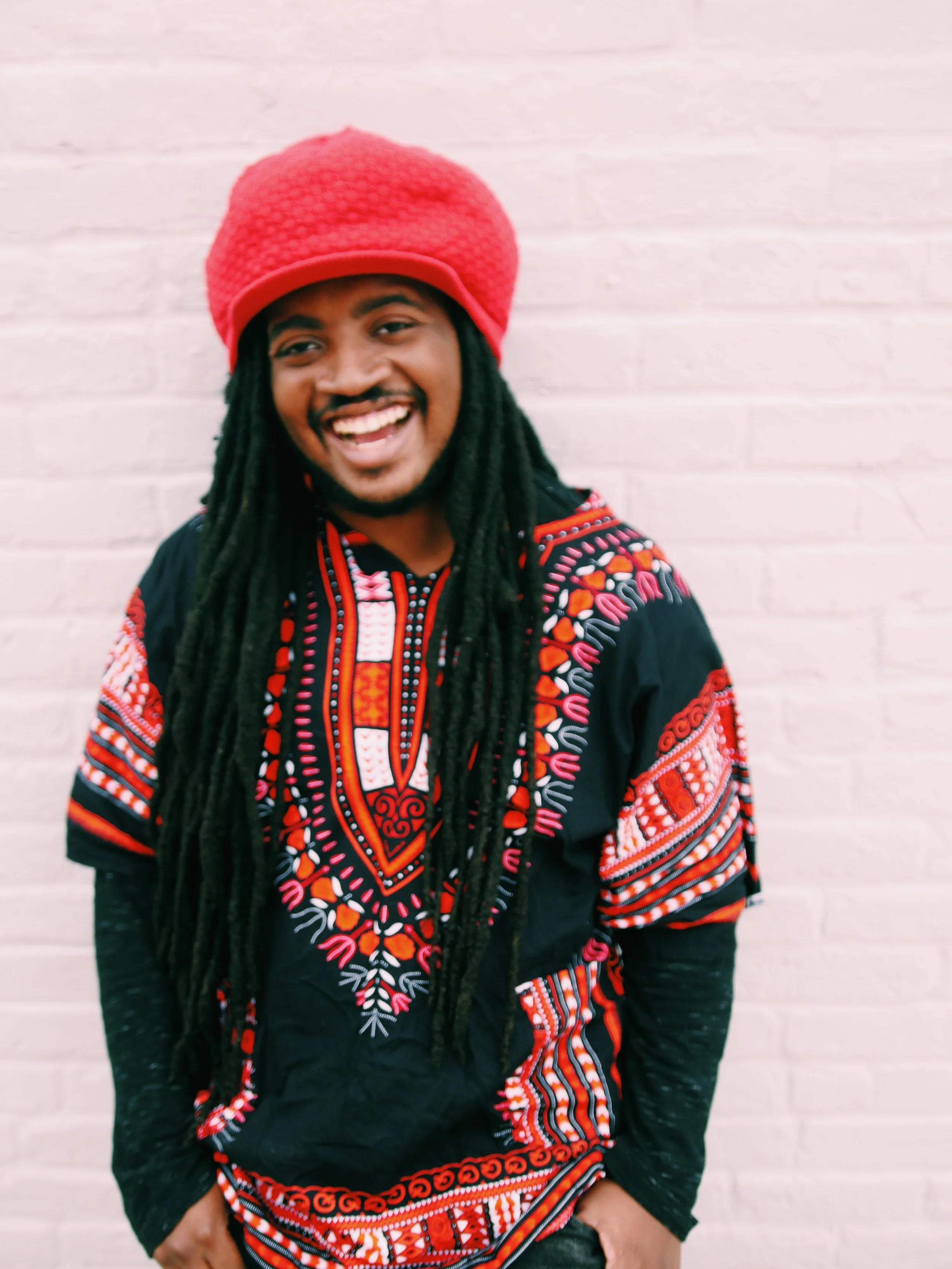 A smiling man with long dreadlocks wearing a bright pink beanie and a black dashiki with red, orange, and white embroidery, standing against a white brick wall.