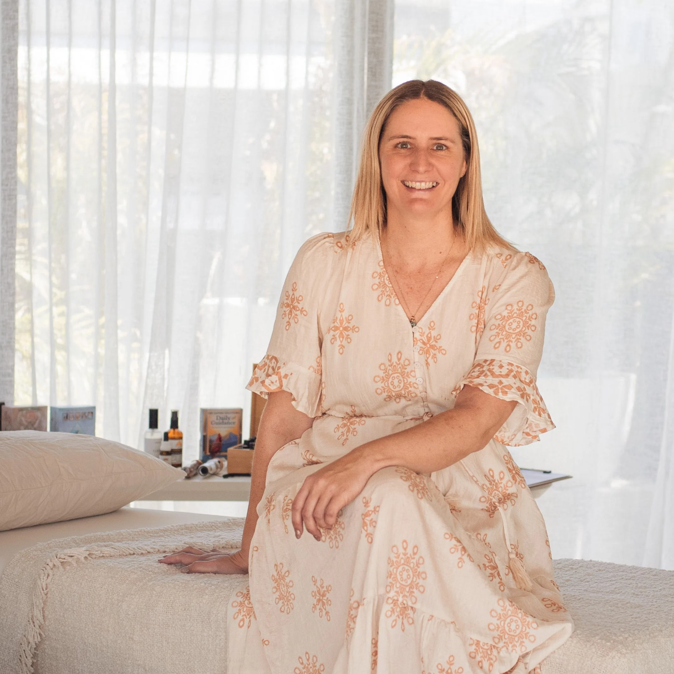 Jackie sits on the edge of a massage table in a room with sheer white curtains and sunlight streaming in.