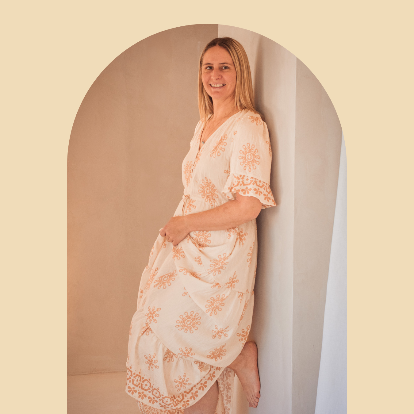Jackie smiling, wearing a cream dress with orange embroidery, standing barefoot in a minimalist room with neutral-colored walls, leaning against an arch.
