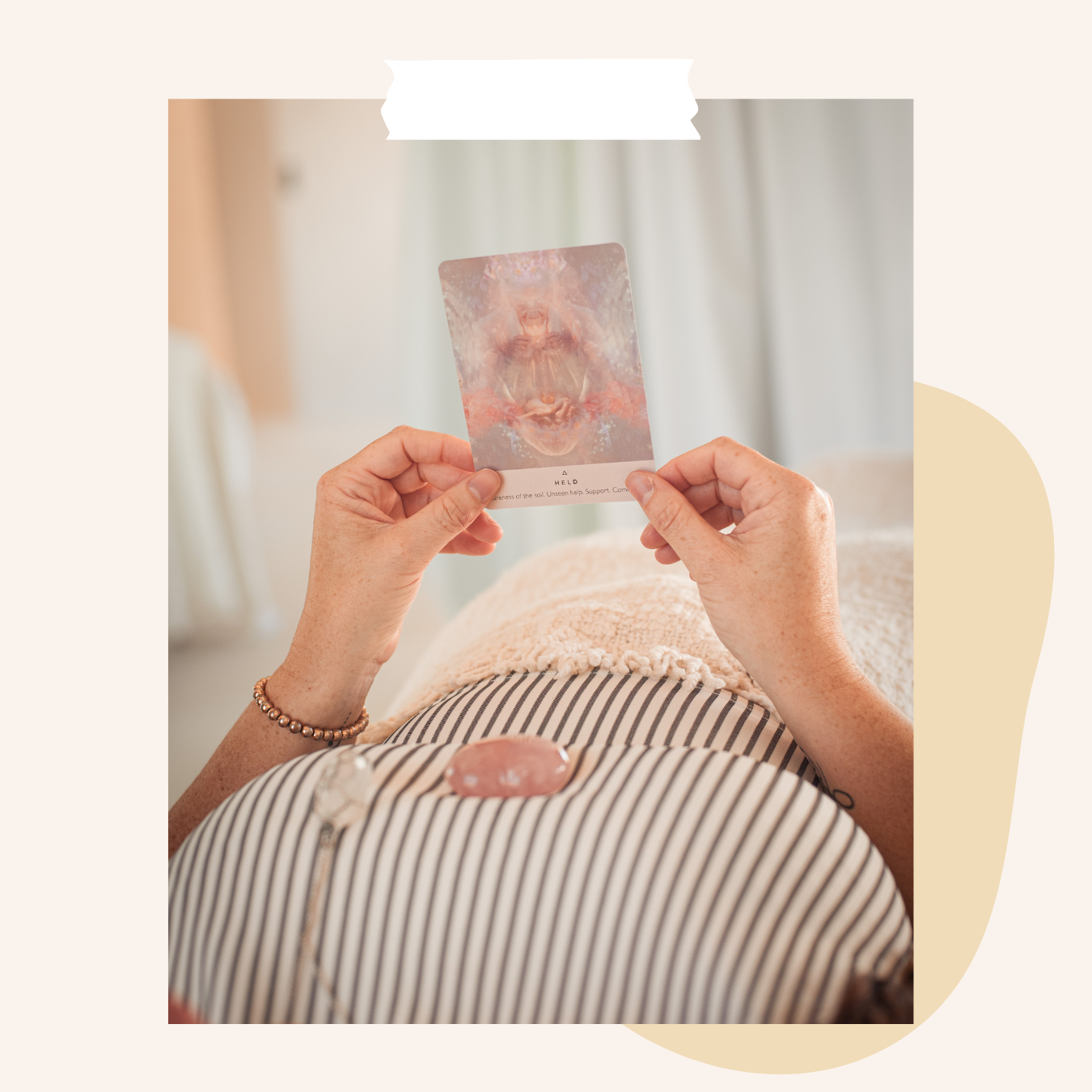 A woman holds an oracle card with spiritual or mystical artwork above their abdomen, which is resting on a striped towel with a rose quartz crystal on their heart chakra.
