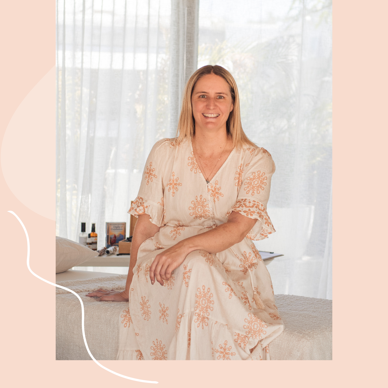 Jackie sits on a massage table in a sunlit room, smiling at the camera, wearing a beige dress with orange embroidery.