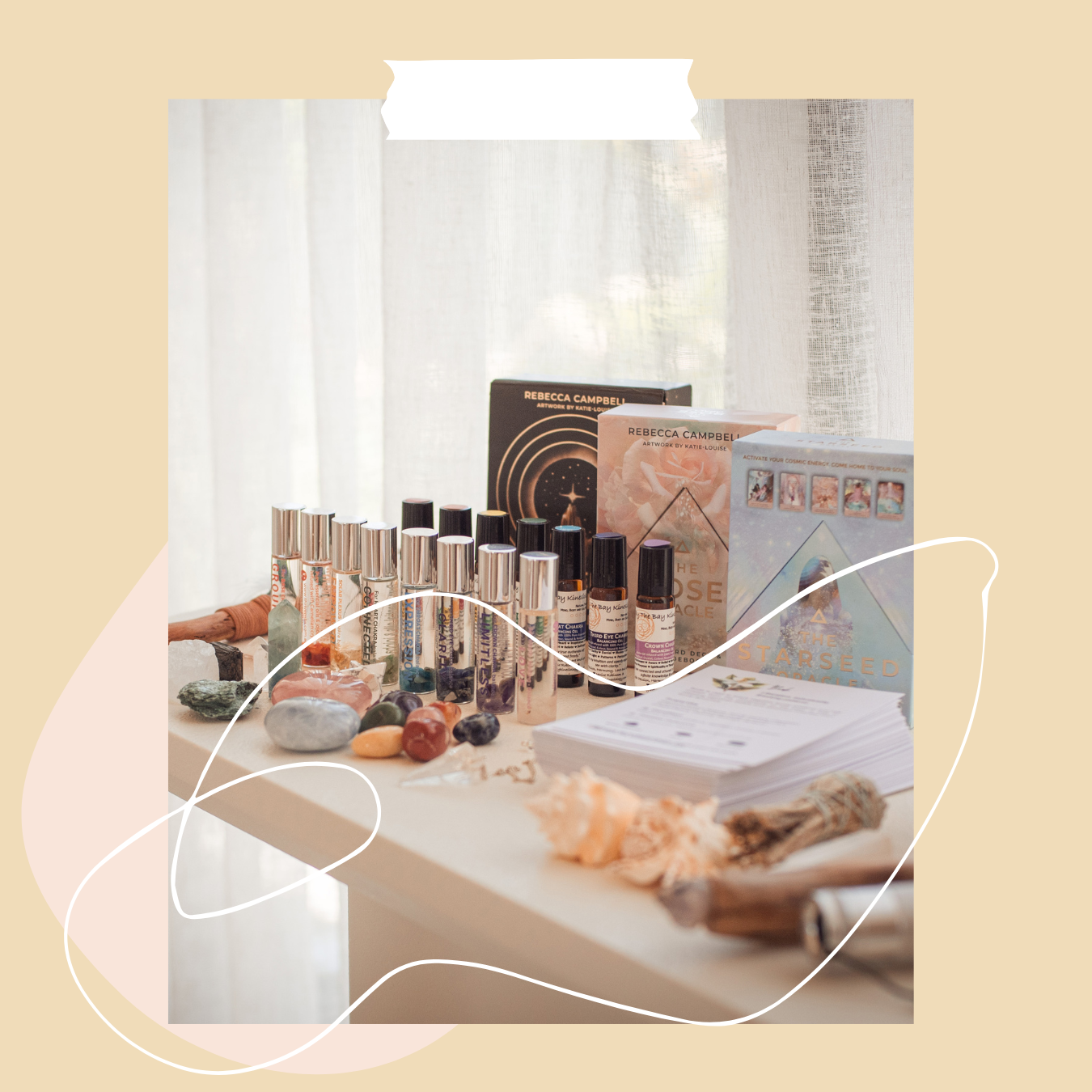 A display of essential oils, oracle cards, crystals, and kinesiology charts on a table near a window with sheer curtains.