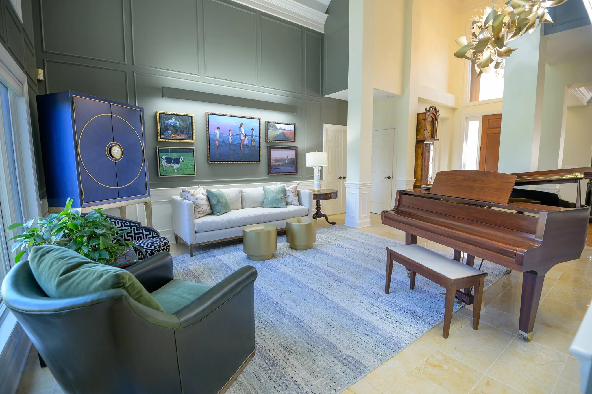 A spacious, well-decorated living room with a green wall adorned with framed artwork, a white sofa with light-colored pillows, a dark blue cabinet with gold accents, a round side table with a lamp, a green armchair, a colorful area rug, and a wooden grand piano with its bench. There is a large window letting in natural light, and a modern chandelier hanging from the ceiling.