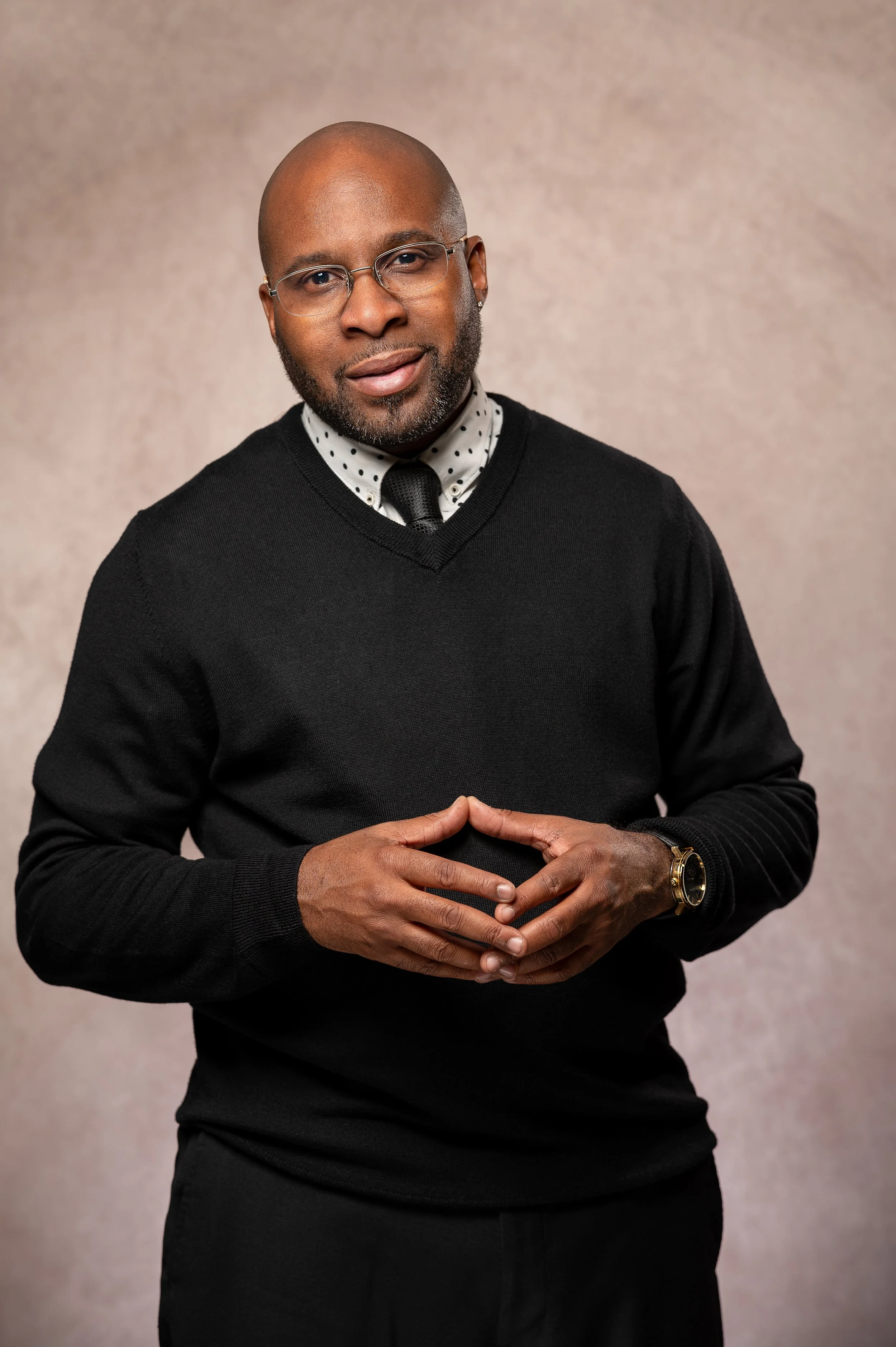 A man with glasses and a beard wearing a black sweater over a polka dot shirt and a black tie, posing with his hands clasped together, against a neutral background.