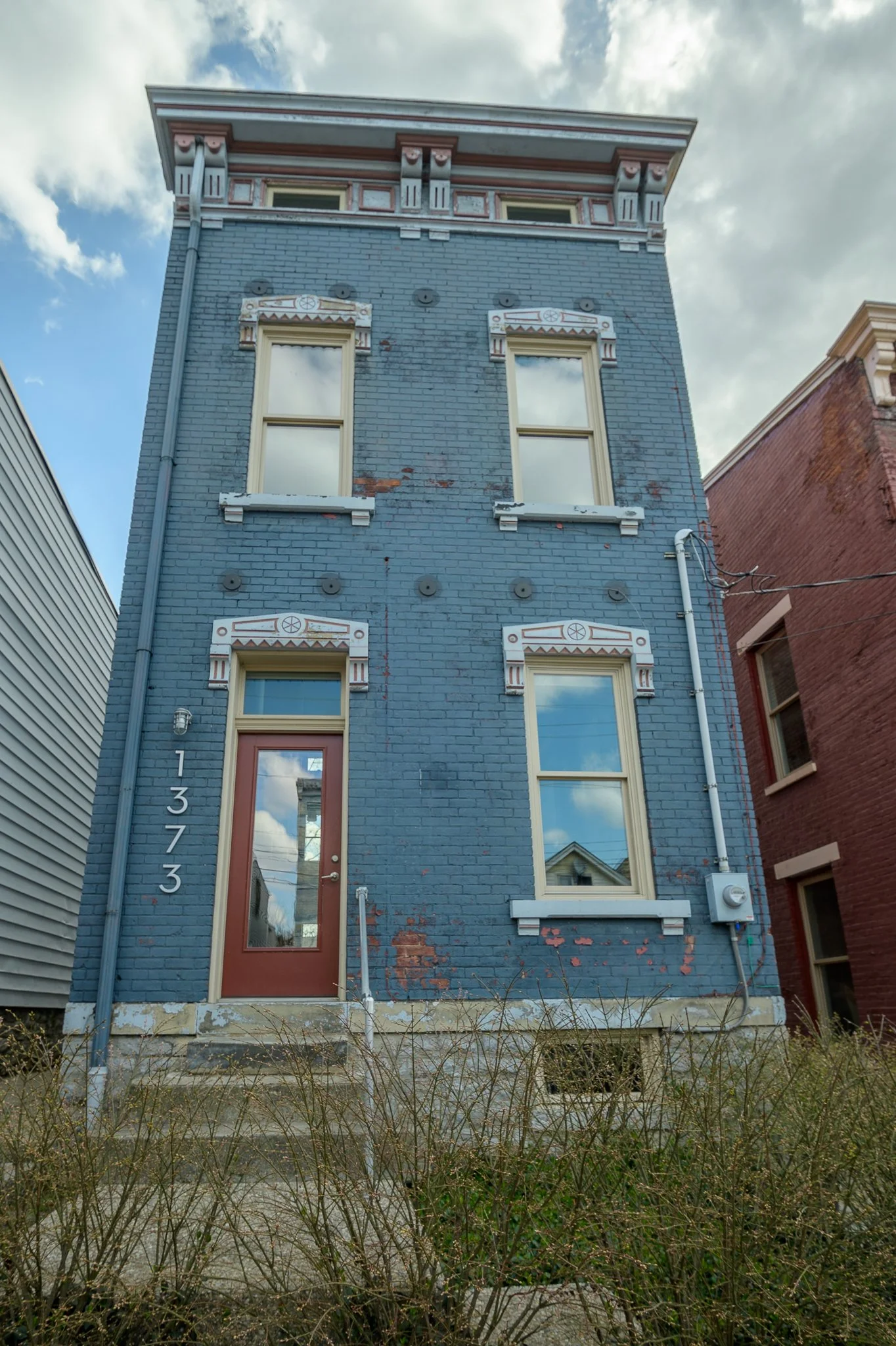 A narrow three-story blue brick house with decorative white trim around the windows and door, located between two other buildings. The house has a brown front door with a window, and the house number 1373 is displayed vertically next to the door. The sky above is partly cloudy.