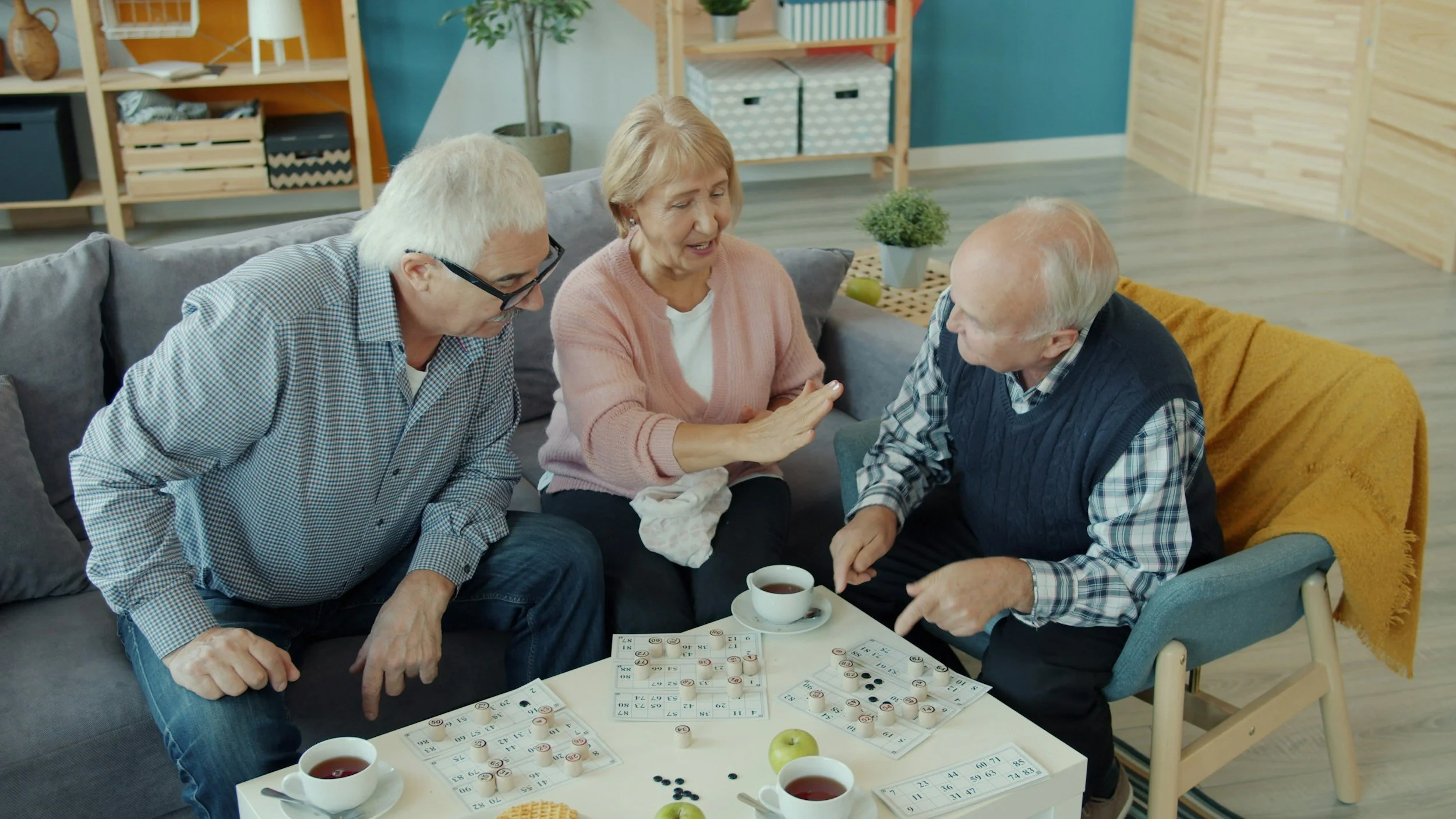Three elderly people sitting around a table playing a tabletop game with bingo cards and chips, drinking tea, and engaging in conversation in a cozy living room.