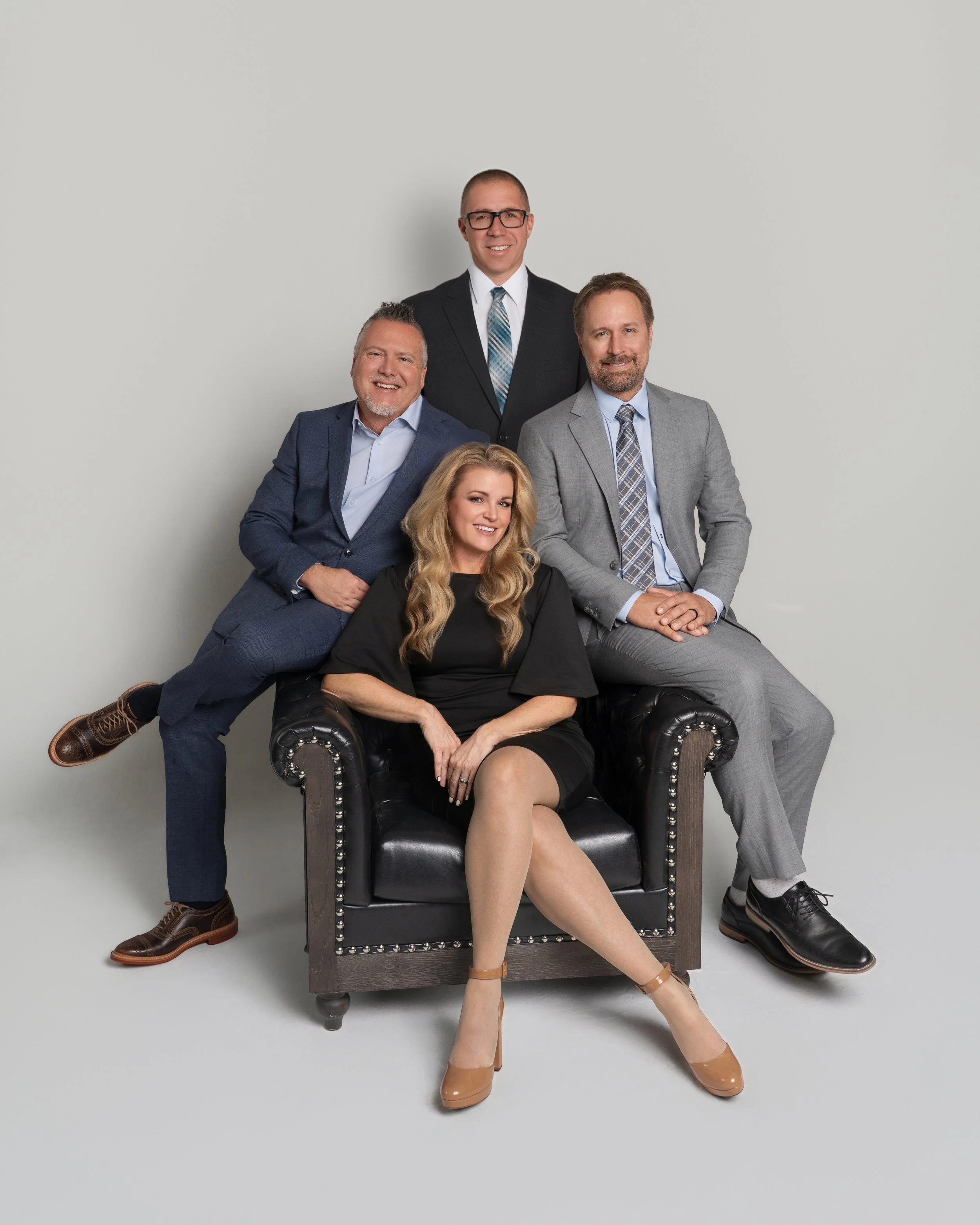 Group of five professionally dressed people posing together in a studio against a plain background, with one woman sitting in a black armchair and four men arranged around her.
