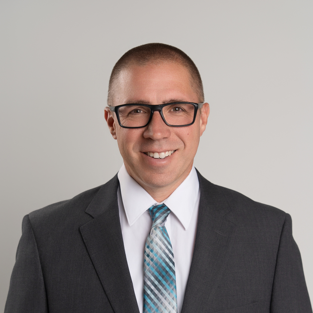 Professional portrait of a man wearing glasses, a white shirt, a gray suit, and a striped tie, smiling against a plain background.