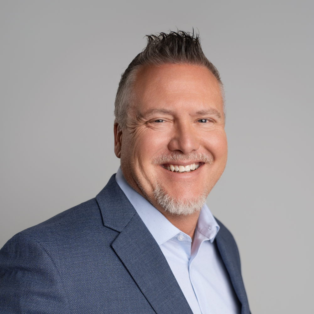 Headshot of a smiling middle-aged man with short, spiked gray hair, a goatee, wearing a navy blazer over a light blue collared shirt, against a plain gray background.