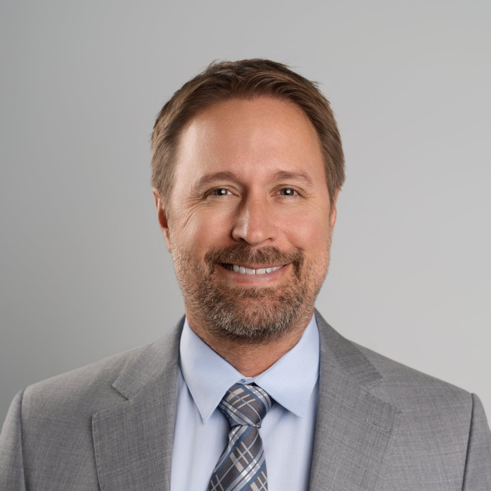 Headshot of a middle-aged man with brown hair and a beard, smiling, wearing a grey suit, light blue shirt, and patterned tie, against a plain grey background.