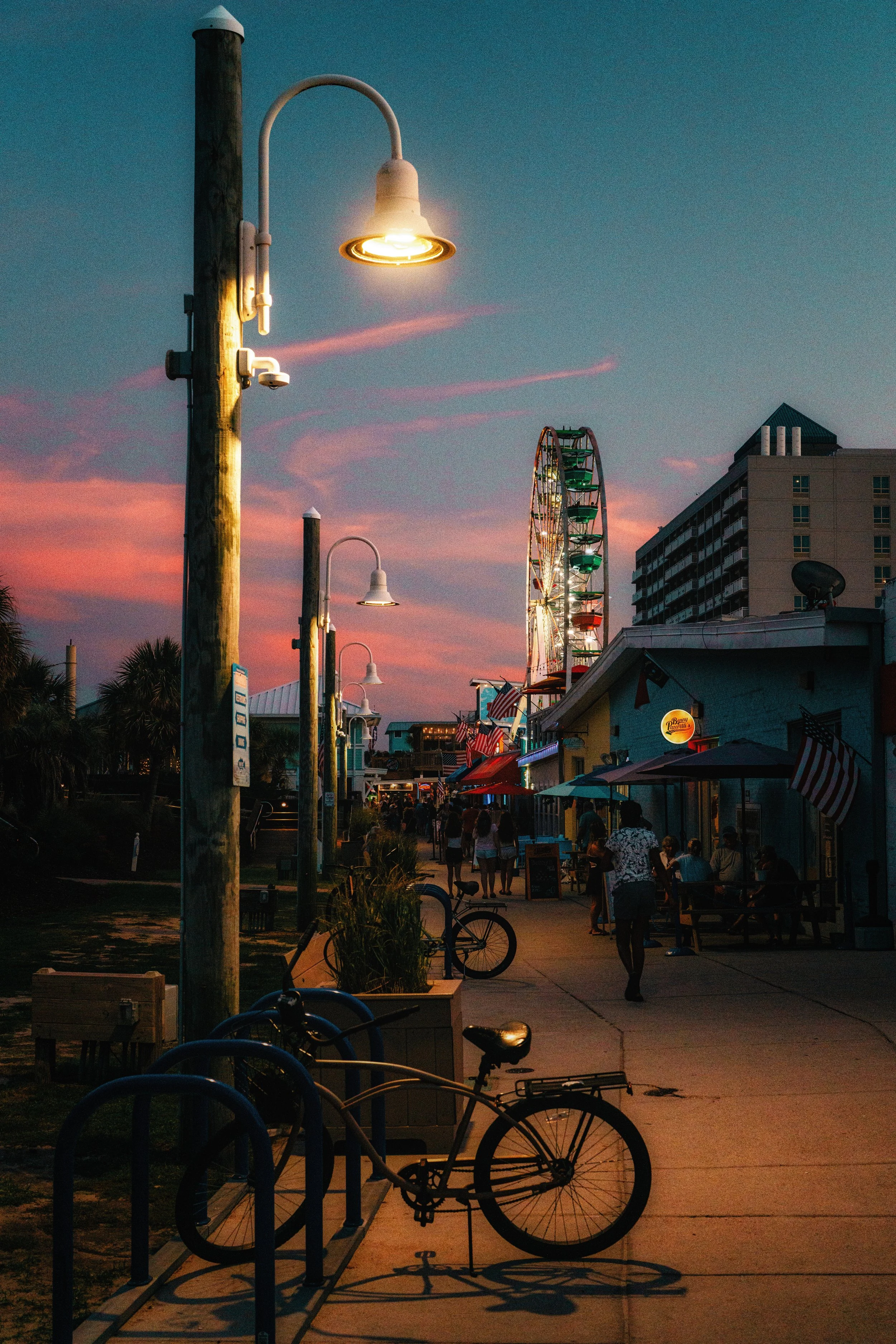 A street scene at dusk with a cloudy sky, an illuminated street lamp, bicycles, and a building with a Ferris wheel in the background.