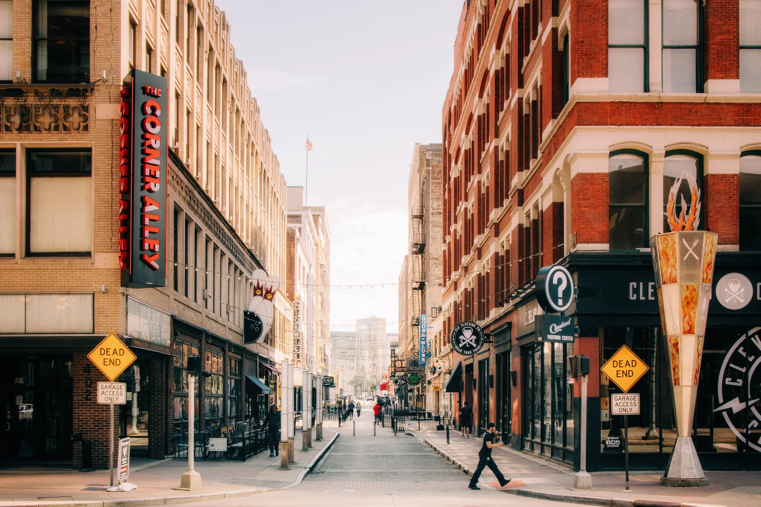 A city street with tall buildings on both sides, storefronts, and signage. Pedestrians are walking along the sidewalks. Signs include a 'Dead End' sign, a 'Garage Access Only' sign, and a marquee for a comedy club. The sky is partly cloudy.