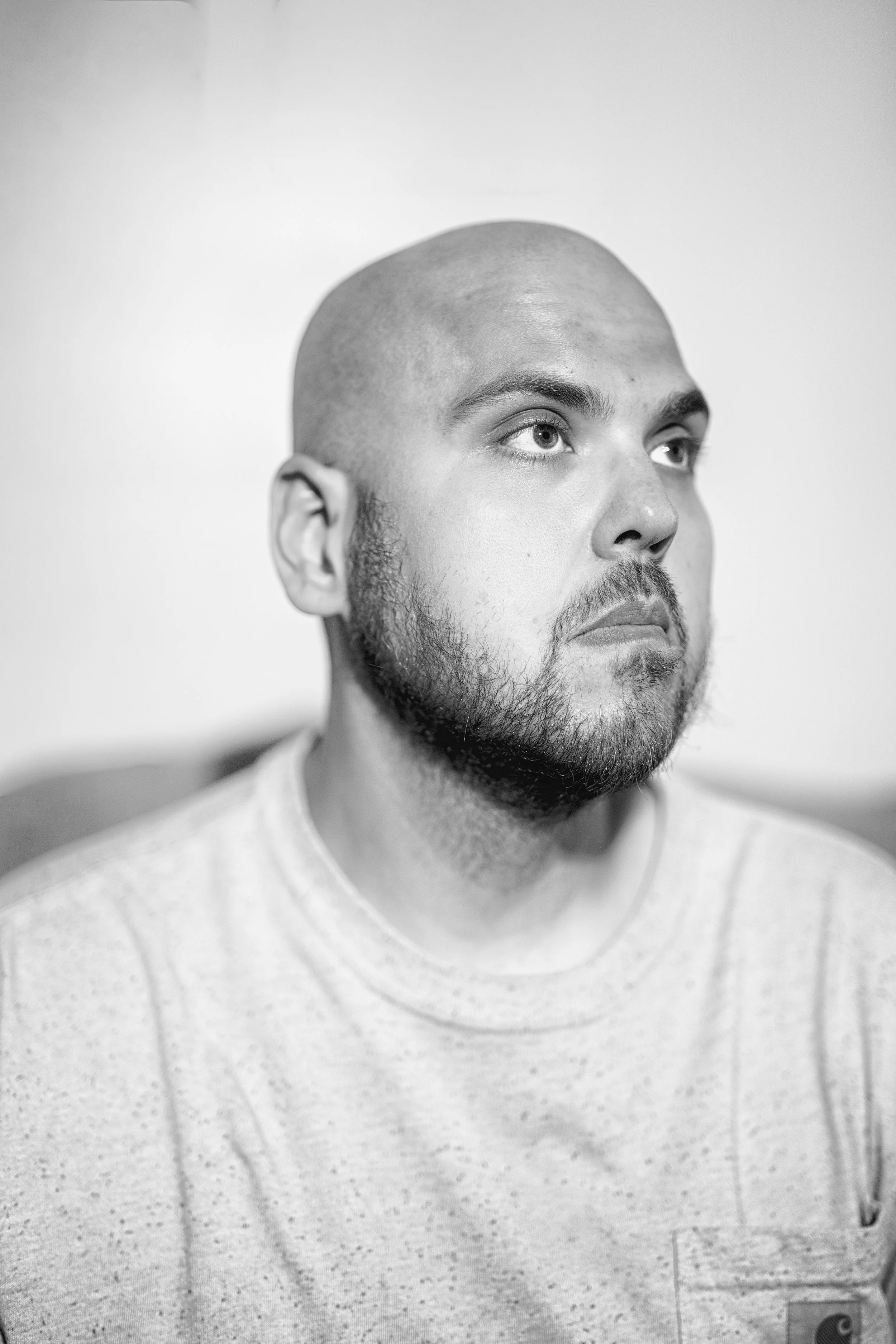 A close-up black-and-white portrait of a man with a shaved head, beard, and mustache, looking slightly to his left, wearing a light-colored t-shirt.
