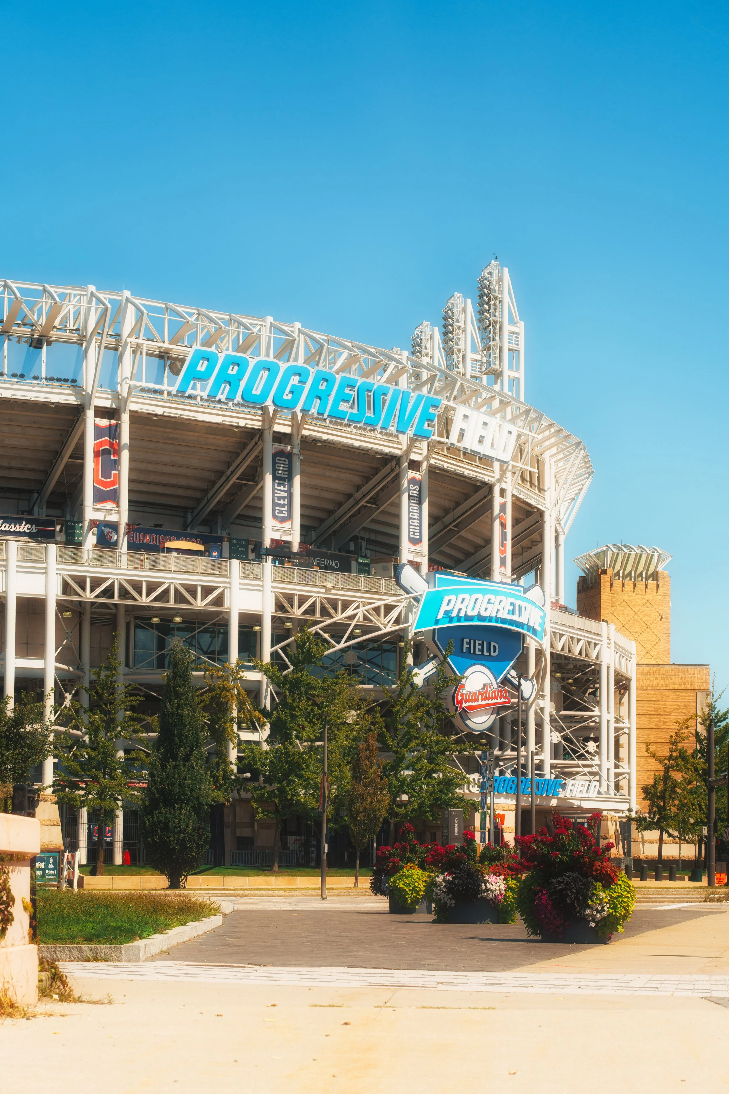 Exterior view of Progressive Field, a Major League Baseball stadium, with blue sky, signage, and landscaped flowerpots.